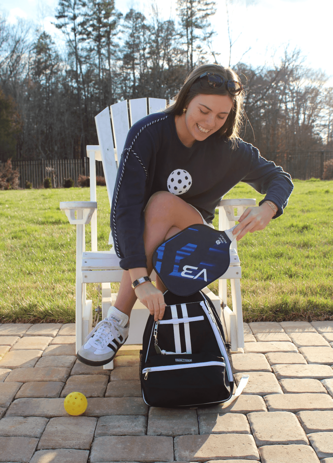 A smiling woman in a navy sweater dress and white sneakers sits outdoors, packing her paddle into the Fresh Pickle Teddi Pickleball Backpack with a roomy interior. A yellow pickleball lies on the patio, with trees and grass in the background.