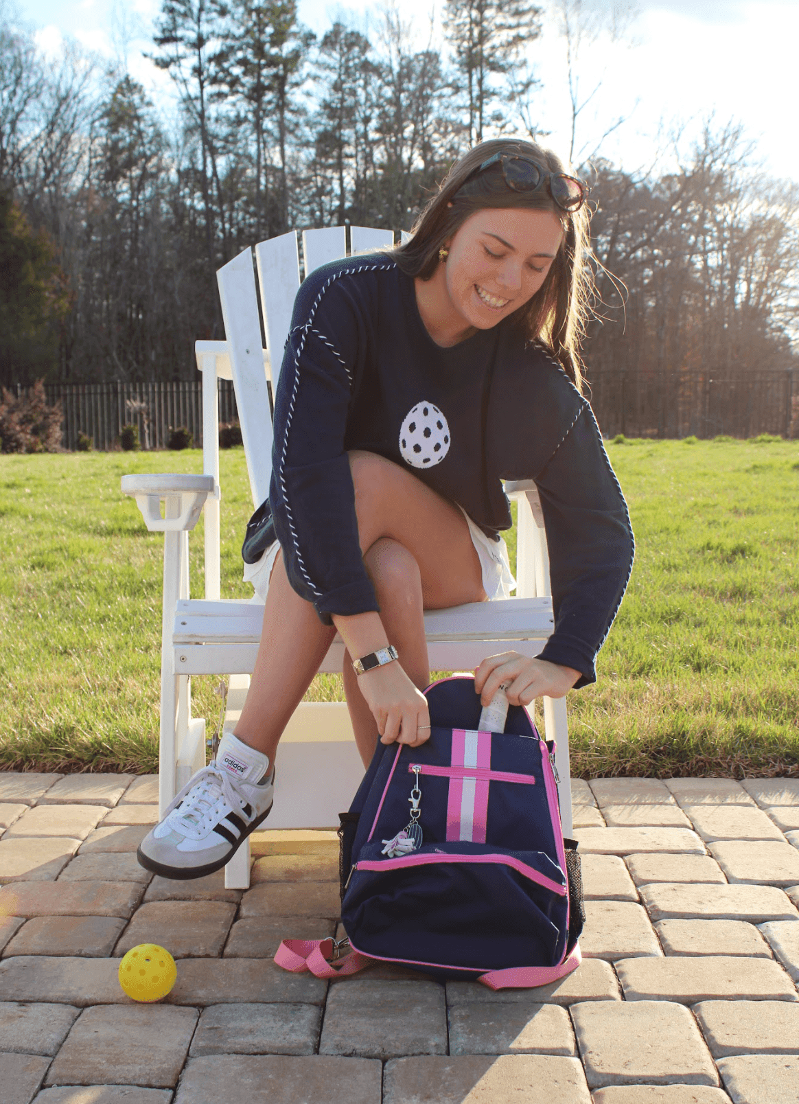A young woman smiles as she unzips her Fresh Pickle Teddi Pickleball Backpack with pink straps while sitting on a white Adirondack chair. She wears a navy shirt with white tennis ball print, a white skirt, and sneakers beside a yellow pickleball.