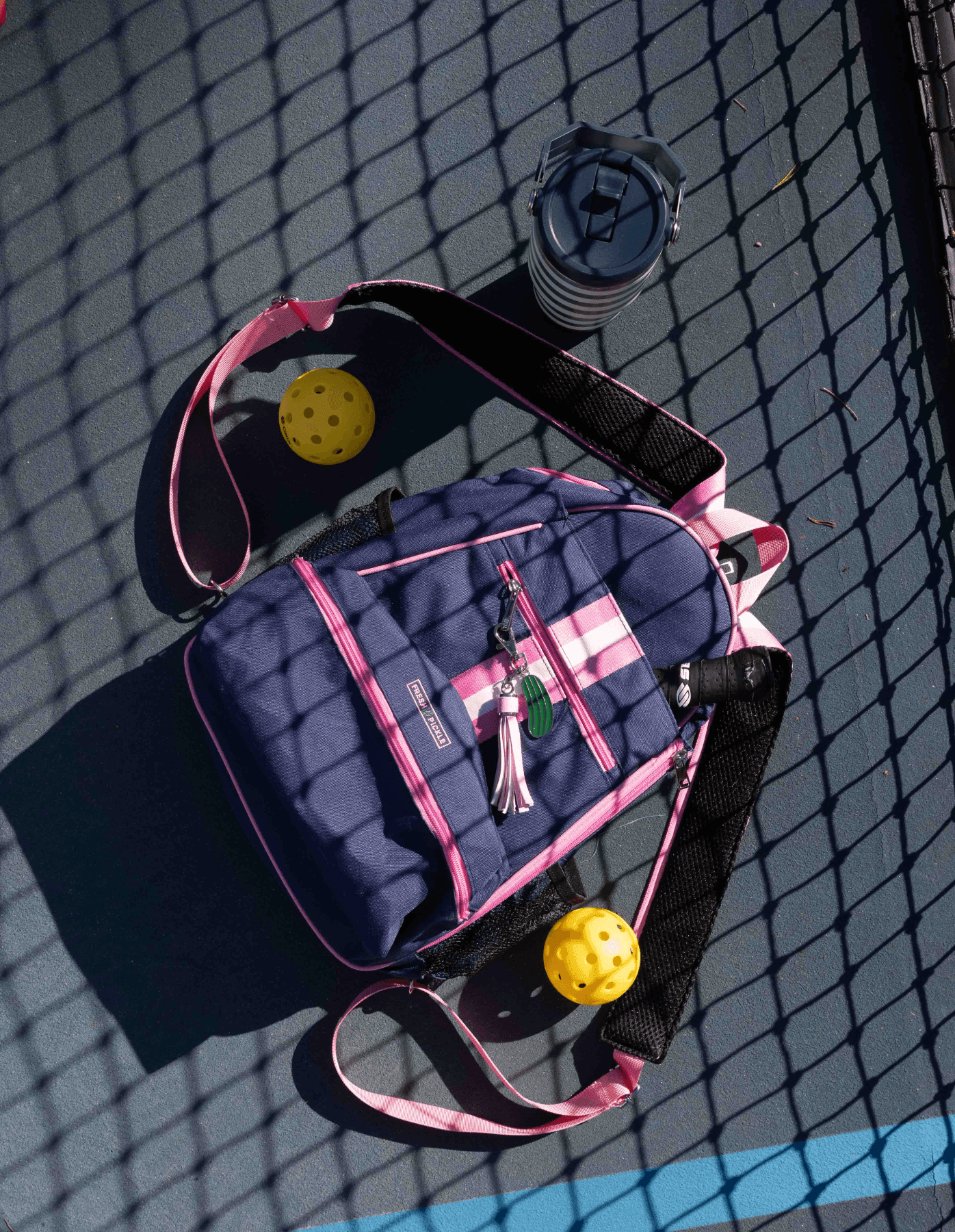 The Fresh Pickle Teddi Pickleball Backpack in blue and pink rests on a sports court, partly shaded by a net. Beside it are two yellow pickleballs, a reusable water bottle, and a keychain, sunlight casting shadows across the scene.