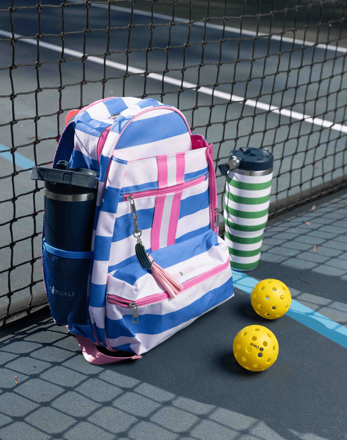 The Fresh Pickle Teddi Pickleball Backpack in blue, white, and pink sits by the net on a tennis court, with two yellow pickleballs and two striped water bottles (green-white and navy-white) nearby.