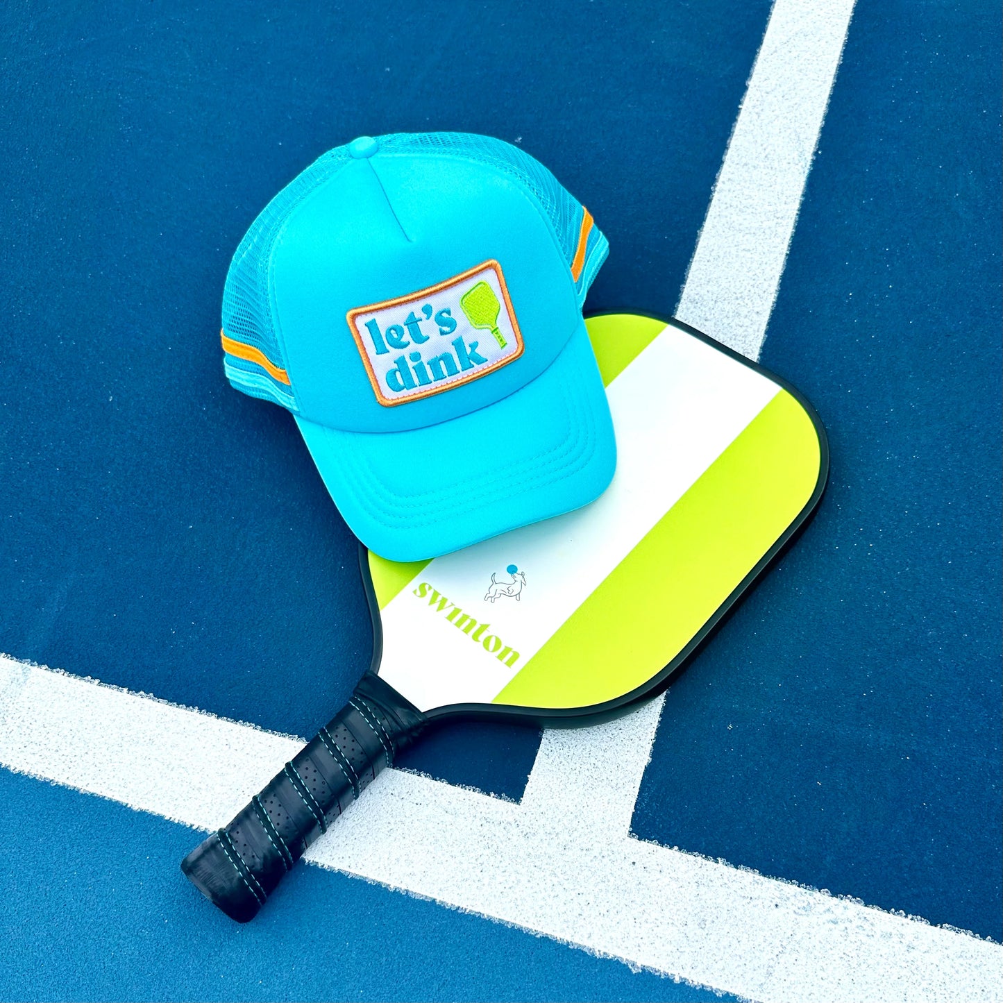 The Swinton Pickleball Dinking Cap Foam Snapback Hat in bright blue with an orange and white "let’s dink" patch, breathable mesh, and adjustable closure sits on a green pickleball paddle beside a white-lined court.