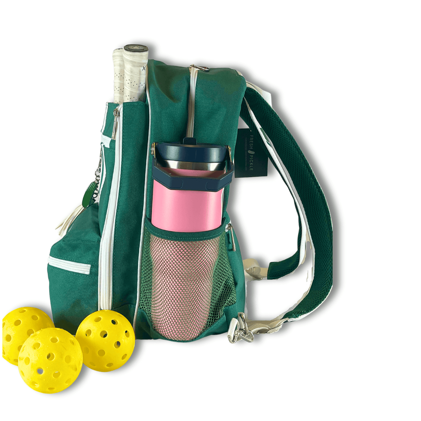 The Fresh Pickle Teddi Pickleball Backpack in green, featuring white zippers and a roomy interior, holds a white-handled racket and pink travel mug in its mesh pocket. Three yellow pickleballs are placed in front on a white background.