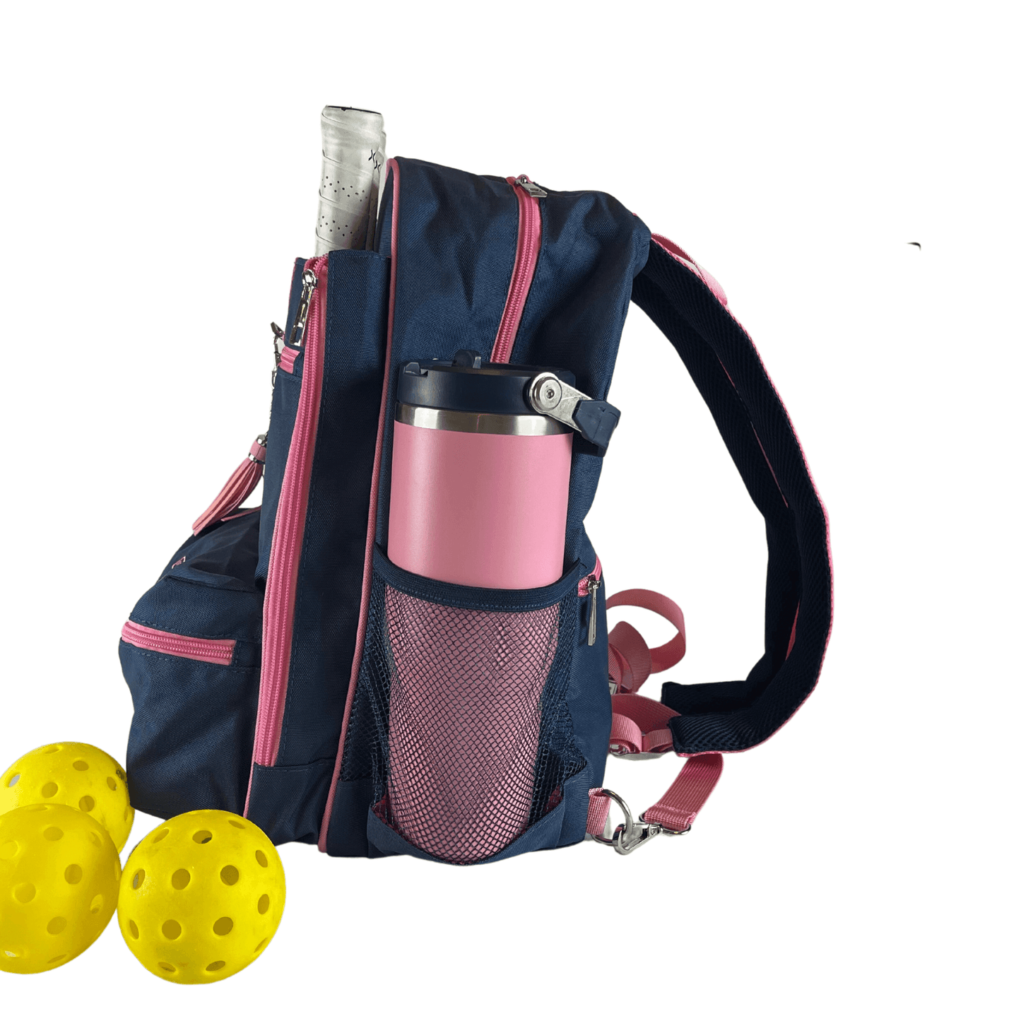 The Fresh Pickle Teddi Pickleball Backpack in navy blue and pink stands upright, carrying a paddle and water bottle with three yellow pickleballs beside it. It features a spacious interior plus mesh and zippered pockets for easy organization.