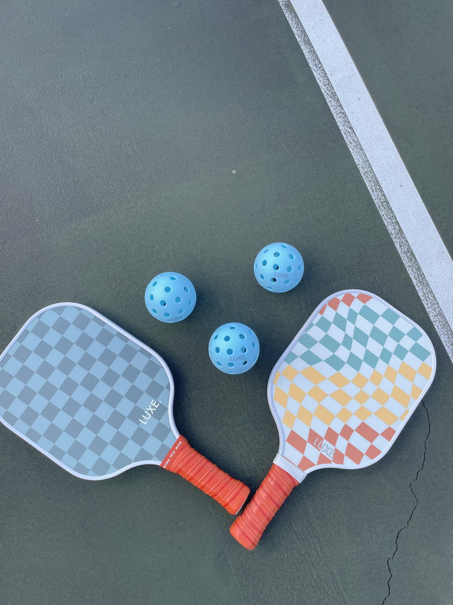 Two Brooke Lynn paddles with checkered designs—one blue-gray, one yellow-blue—are on a court near a white line. Above them are three LUXE Pickleball Sky Pickleballs (outdoor, blue, pack of 3).
