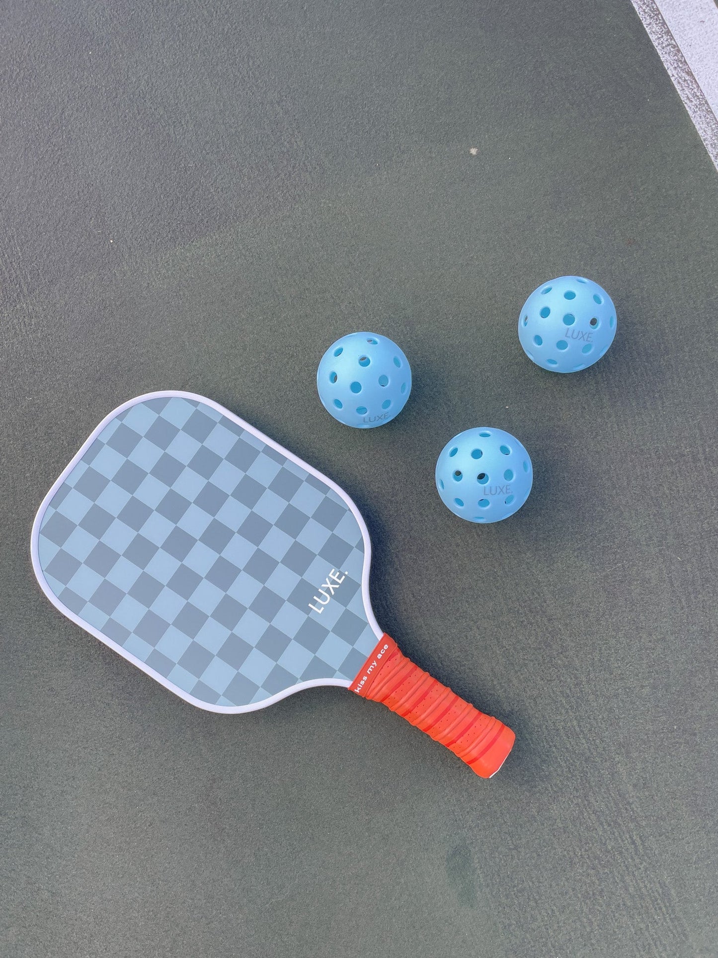 A Brooke Lynn paddle with a checkered pattern and red handle lies on a green court next to three LUXE Pickleball Sky Pickleballs.