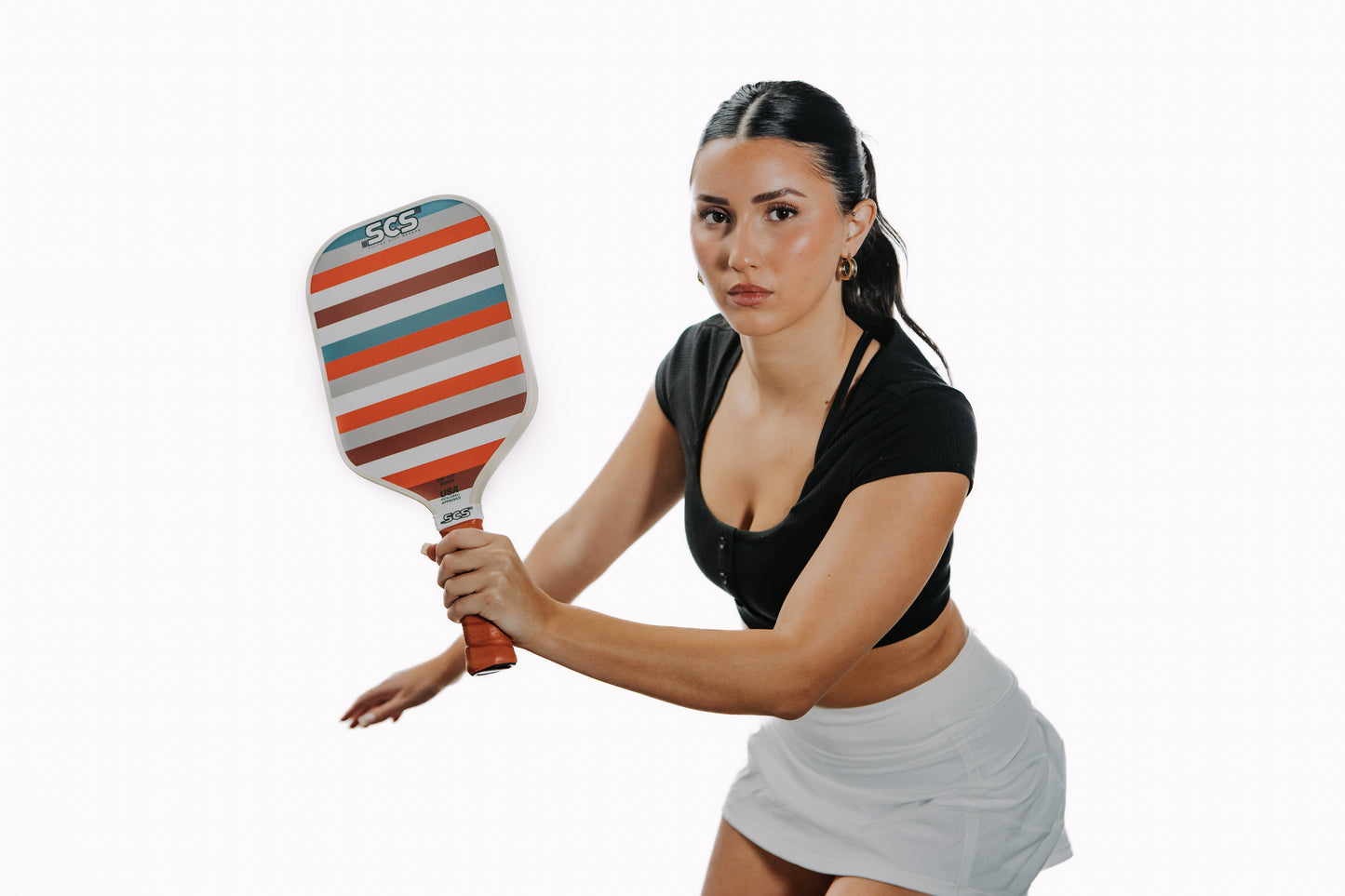 A focused woman with dark hair pulled back, wearing a black top and white skirt, holds the Salted City Sports Slade | Evo-Lite Series pickleball paddle in an athletic stance against a plain white background.