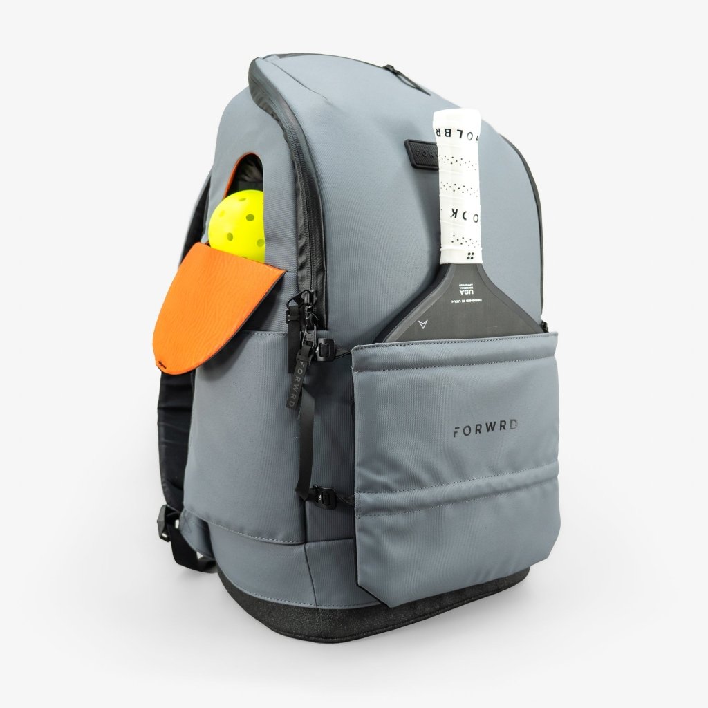 A gray FORWRD Court Caddy Front Paddle Sleeve backpack stands upright with a front pocket, an orange cap and yellow ball in a side pocket, and a black paddle partly tucked into the main compartment, all on a white background.