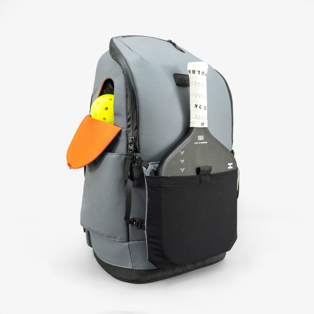 A FORWRD Court Caddy Front Paddle Sleeve is attached to the front of a gray backpack with a black bottom, shown against a white background. An orange cap, yellow ball in the side pocket, and a white luggage tag complete the look.