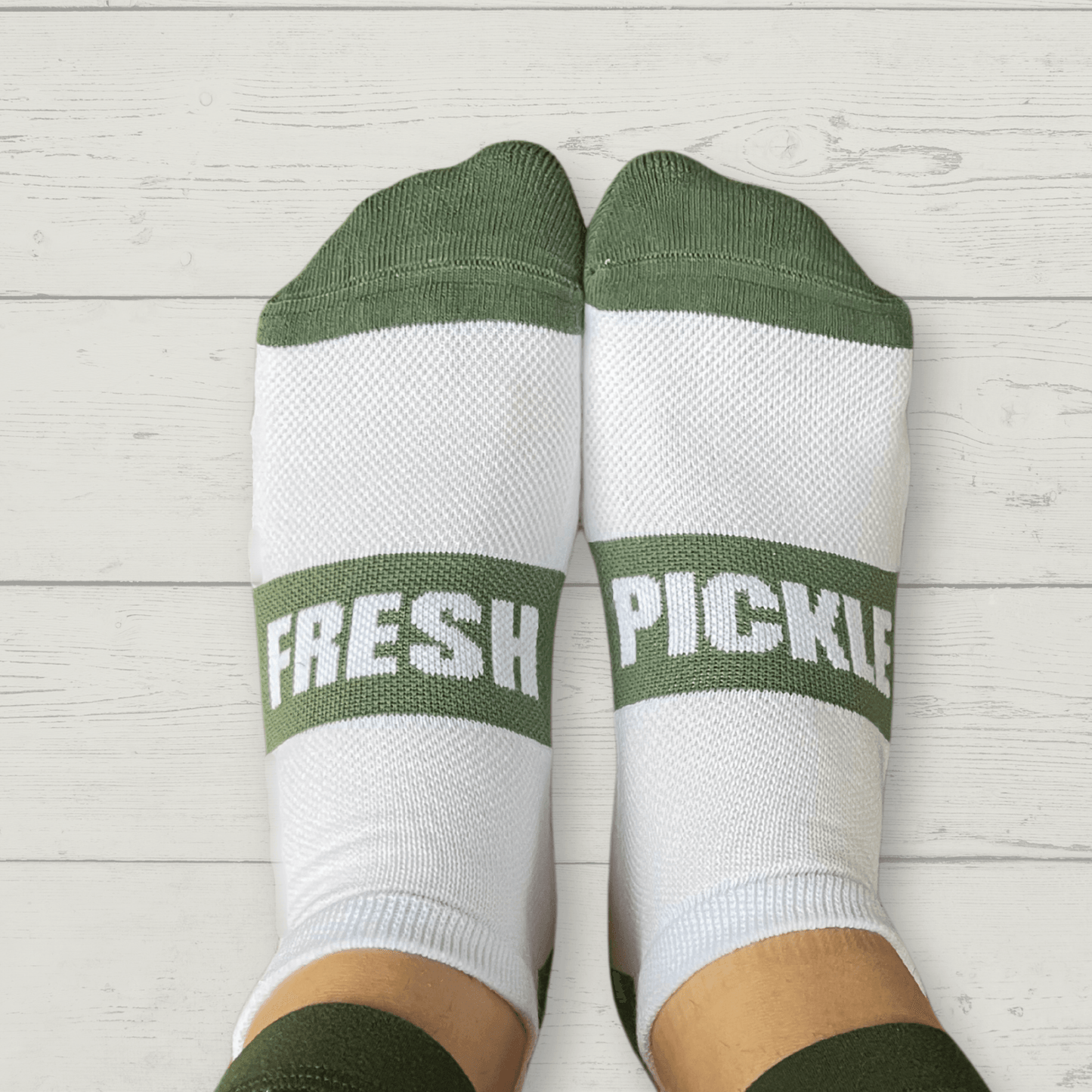 A pair of feet on a white wooden floor wear Fresh Pickle brand Pickleball Socks—breathable white socks with green toes, heels, and cuffs. "FRESH" is on the left sock and "PICKLE" on the right in bold green letters.
