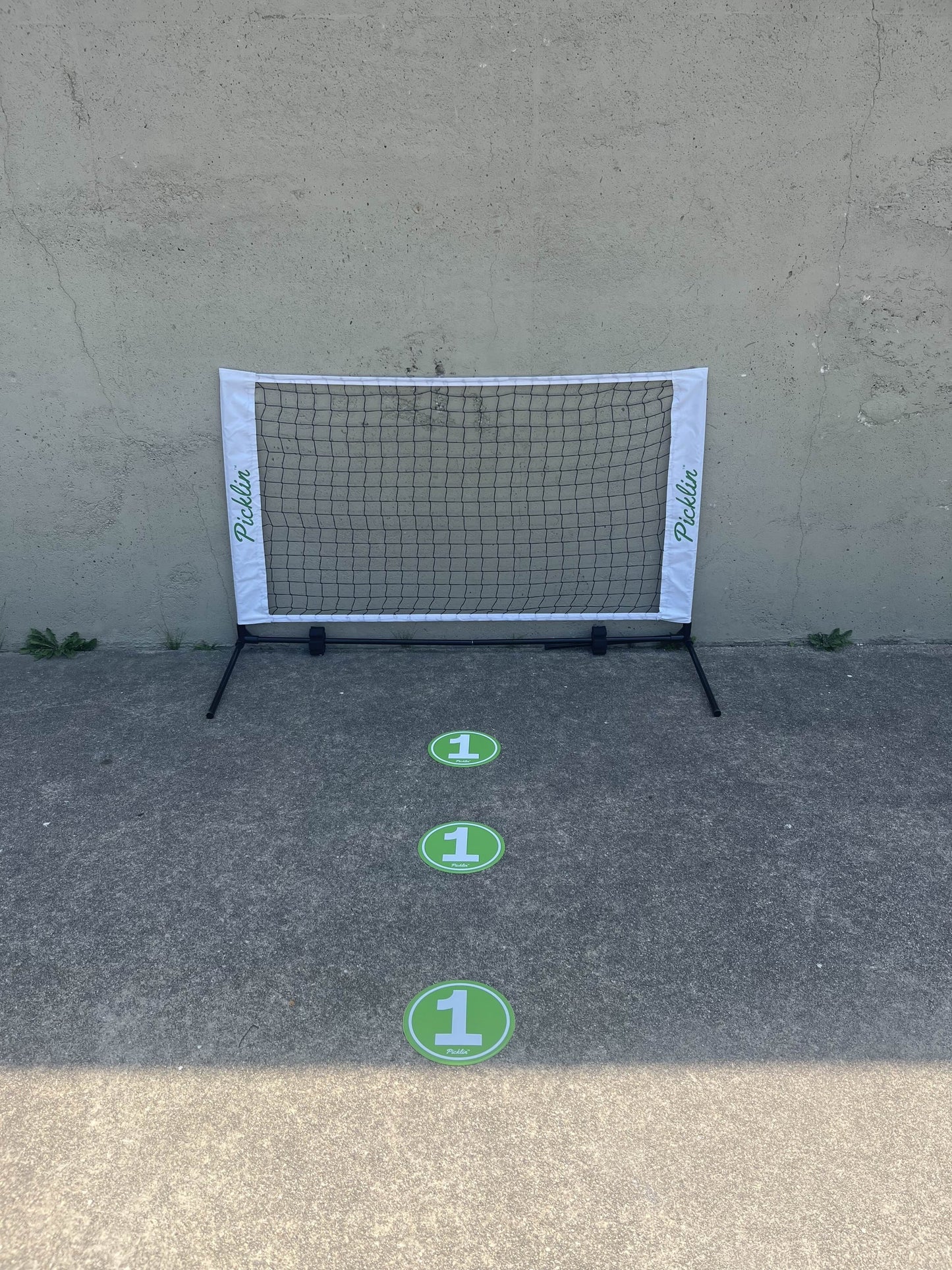 A Picklin™ Dink Net with a white Paddle frame stands by a wall. Three green Picklin™ Floor Targets, each marked with a large white "1," are lined up on the ground, leading to the net for training.