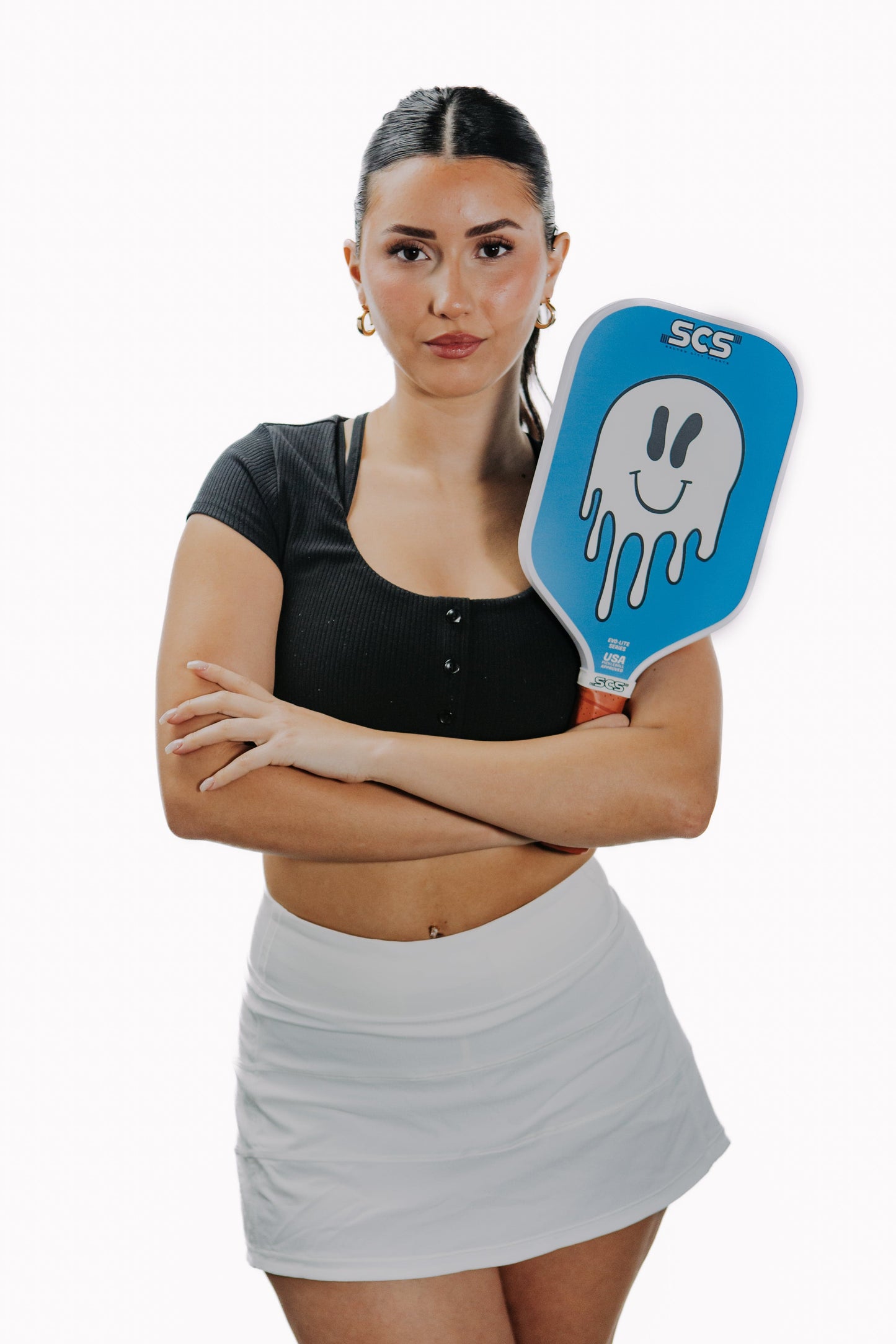 A woman with long dark hair in a ponytail stands confidently, arms crossed, holding the Salted City Sports Sonrisa Evo-Lite Series pickleball paddle with a smiling dripping ghost graphic. She wears hoop earrings, a black crop top, and a white tennis skirt.