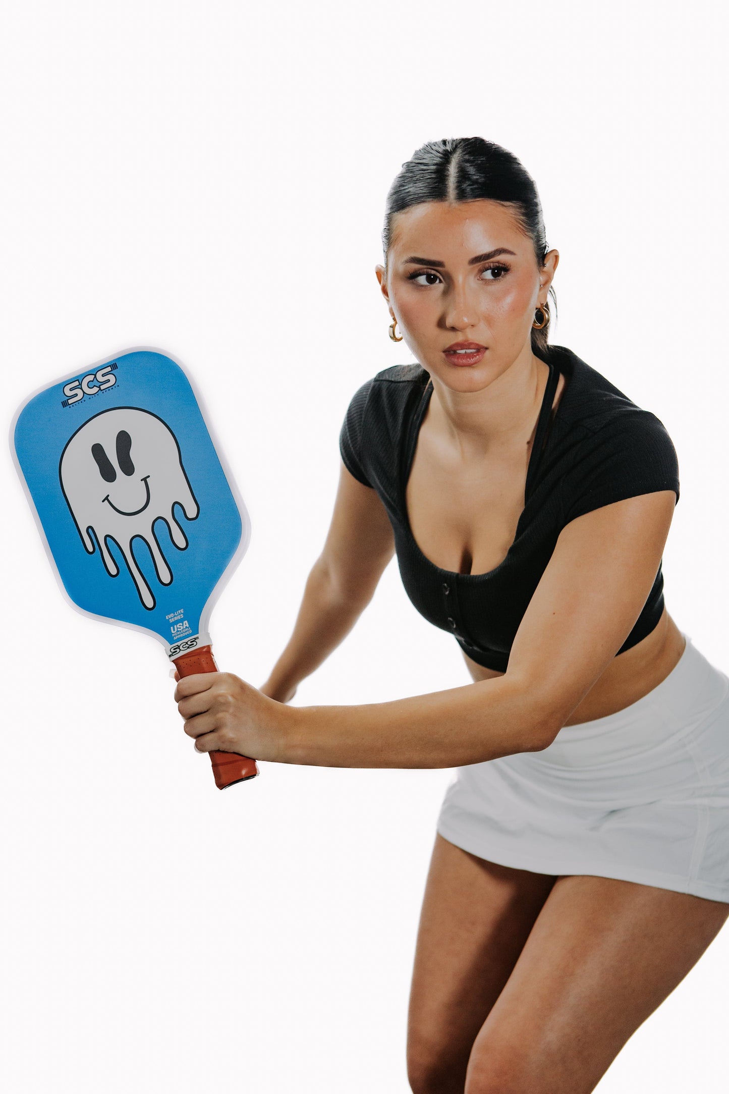 A woman with dark hair tied back, wearing a black crop top and white skirt, holds the Salted City Sports Sonrisa Evo-Lite Series pickleball paddle, featuring a smiling, melting face design. She stands in an athletic pose against a white background.