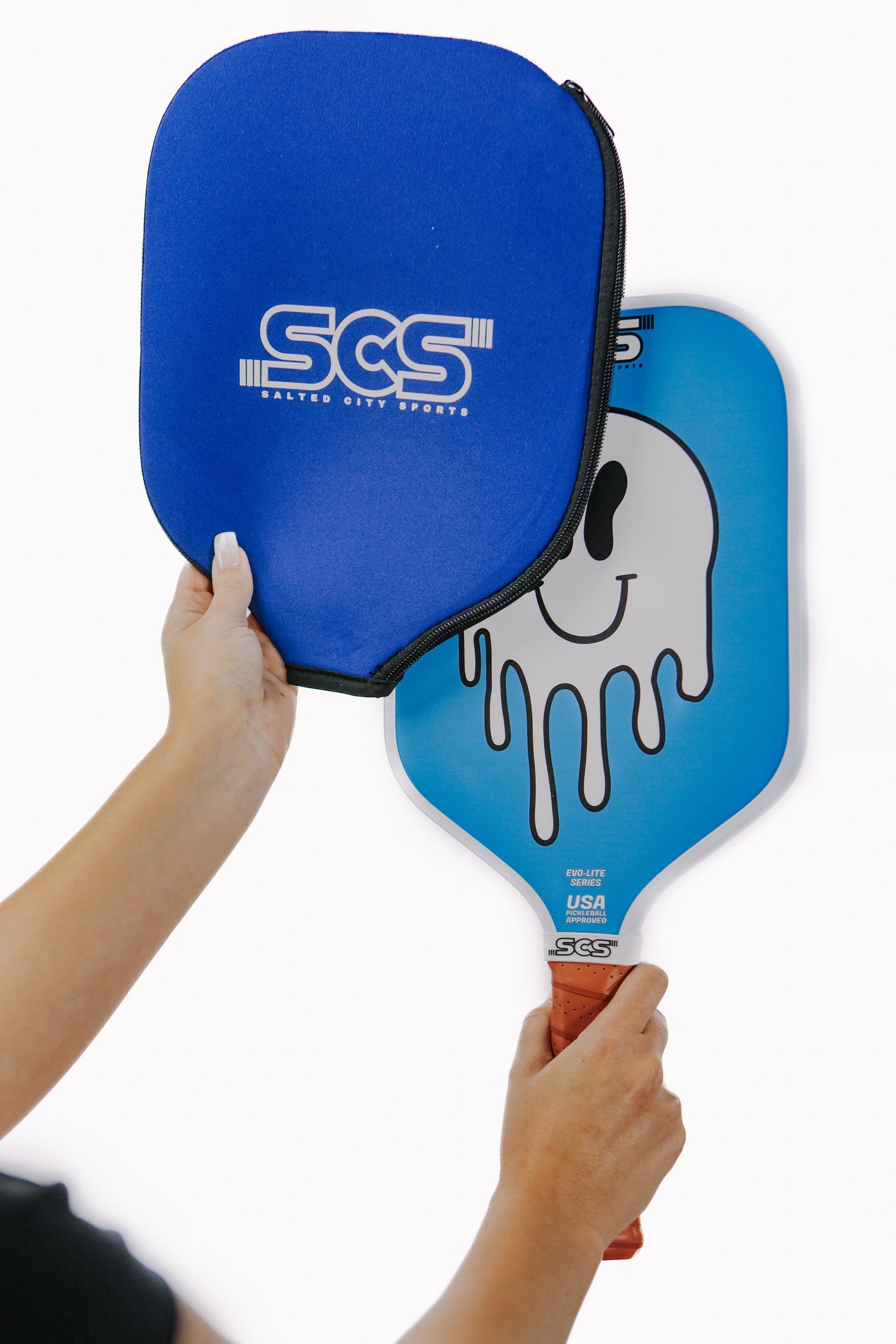 A person puts a blue Salted City Sports SCS paddle cover on the Sonrisa | Evo-Lite Series paddle, which features a white melting smiley face design and brown grip. Both are USA Pickleball approved and shown against a plain white background.