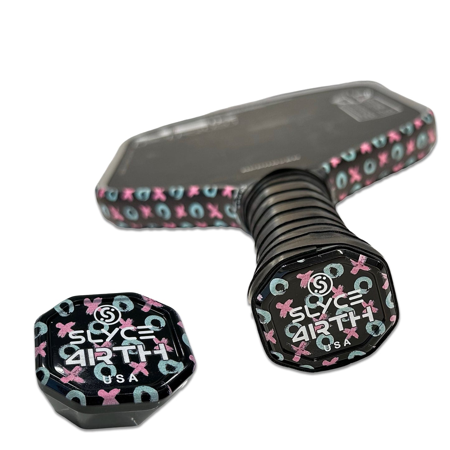 A close-up of two Slyce Sport Special Edition 4rth XO’s SpeedCap (28g, 1.0 oz.) Paddle Performance Enhancers—both black with blue and pink XO symbols. One cap is on the handle, the other nearby; the paddle edge features a matching pattern.