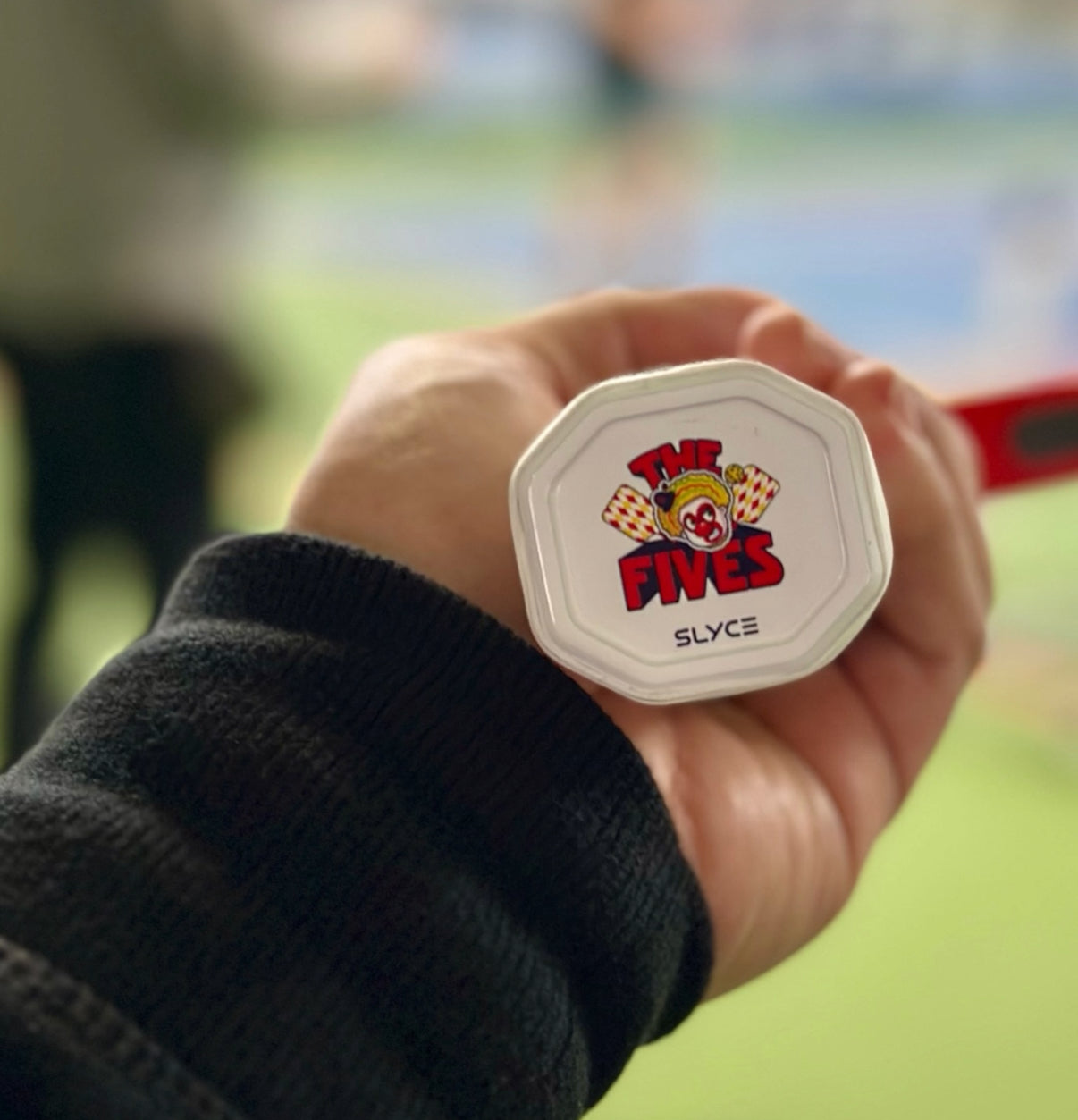 A hand grips the Slyce Sport Special Edition New Jersey Five’s SpeedCap (28g, 1.0 oz.) Paddle Performance Enhancer - Purple, featuring “THE FIVES” logo with two clown faces; a blurred green pickleball court is in the background.