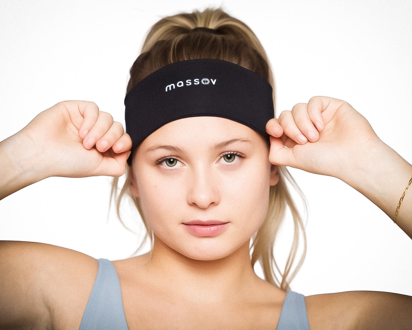 A young blonde woman with light skin wears a Massov Sports Headband in black, facing forward and holding it with both hands. She is dressed in a light blue sleeveless top against a plain white background.