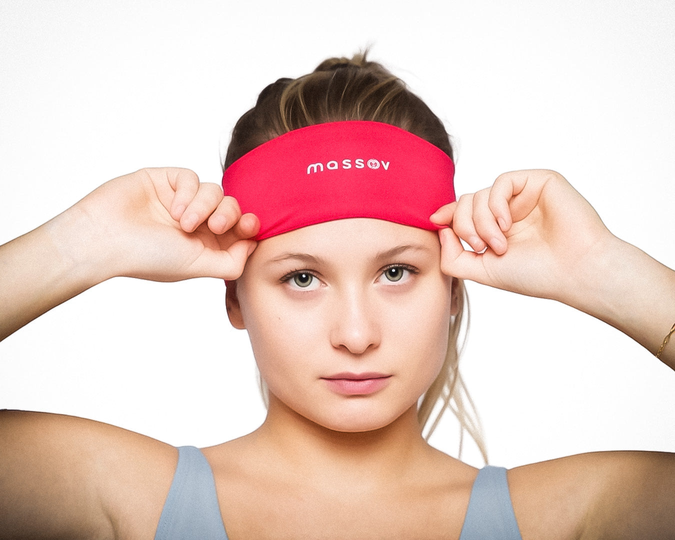 A young woman with light skin and green eyes adjusts a bright red Massov Sports Headband, her light brown hair pulled back. She wears a light blue tank top and looks directly at the camera against a plain white background.