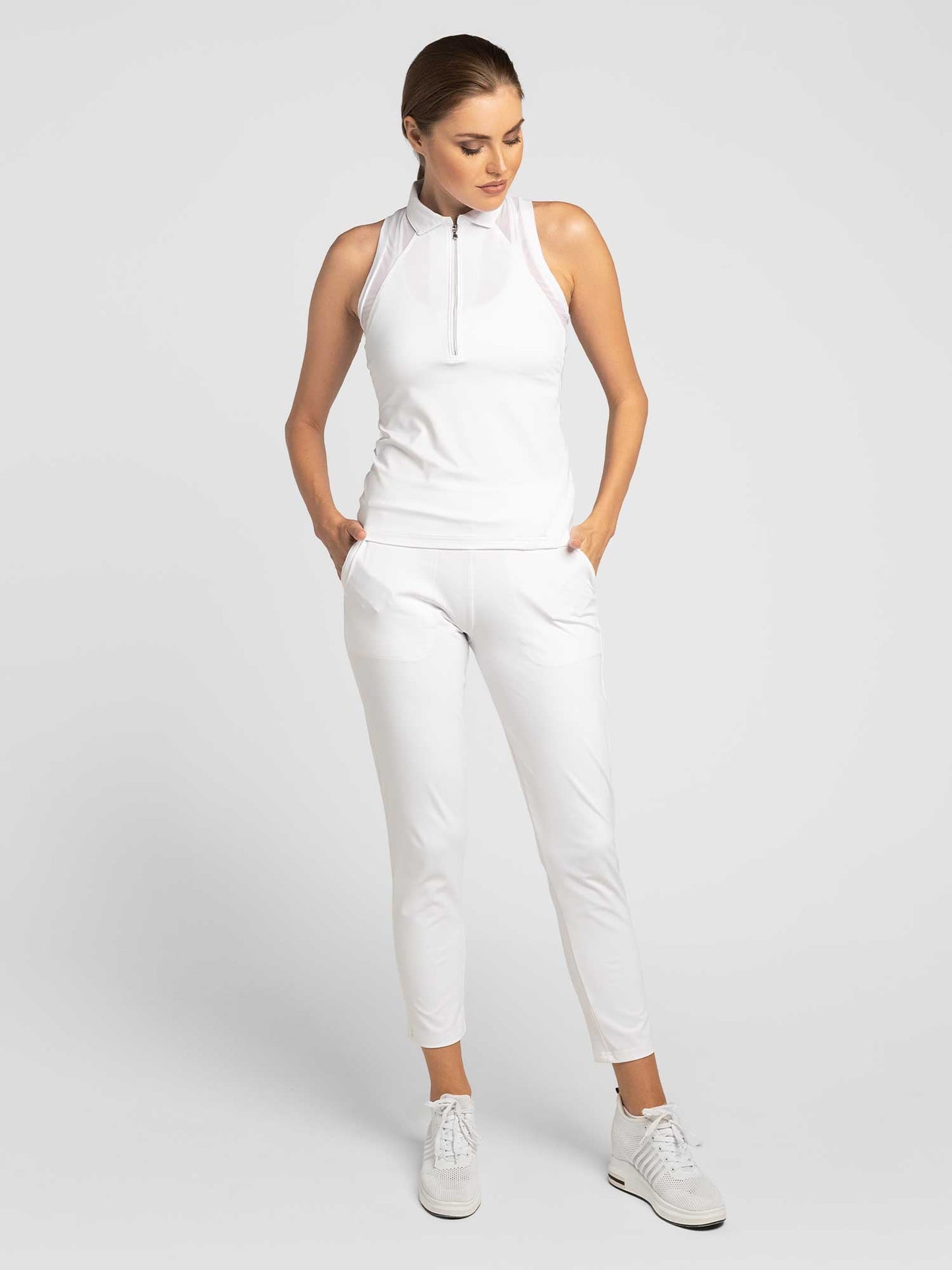 A woman with short brown hair stands against a light background, hands in pockets, wearing inPhorm NYC's Stylish Women's Golf Pants—white, fitted, and moisture-wicking—with a sleeveless white zip-up top. She gazes down and to the side.