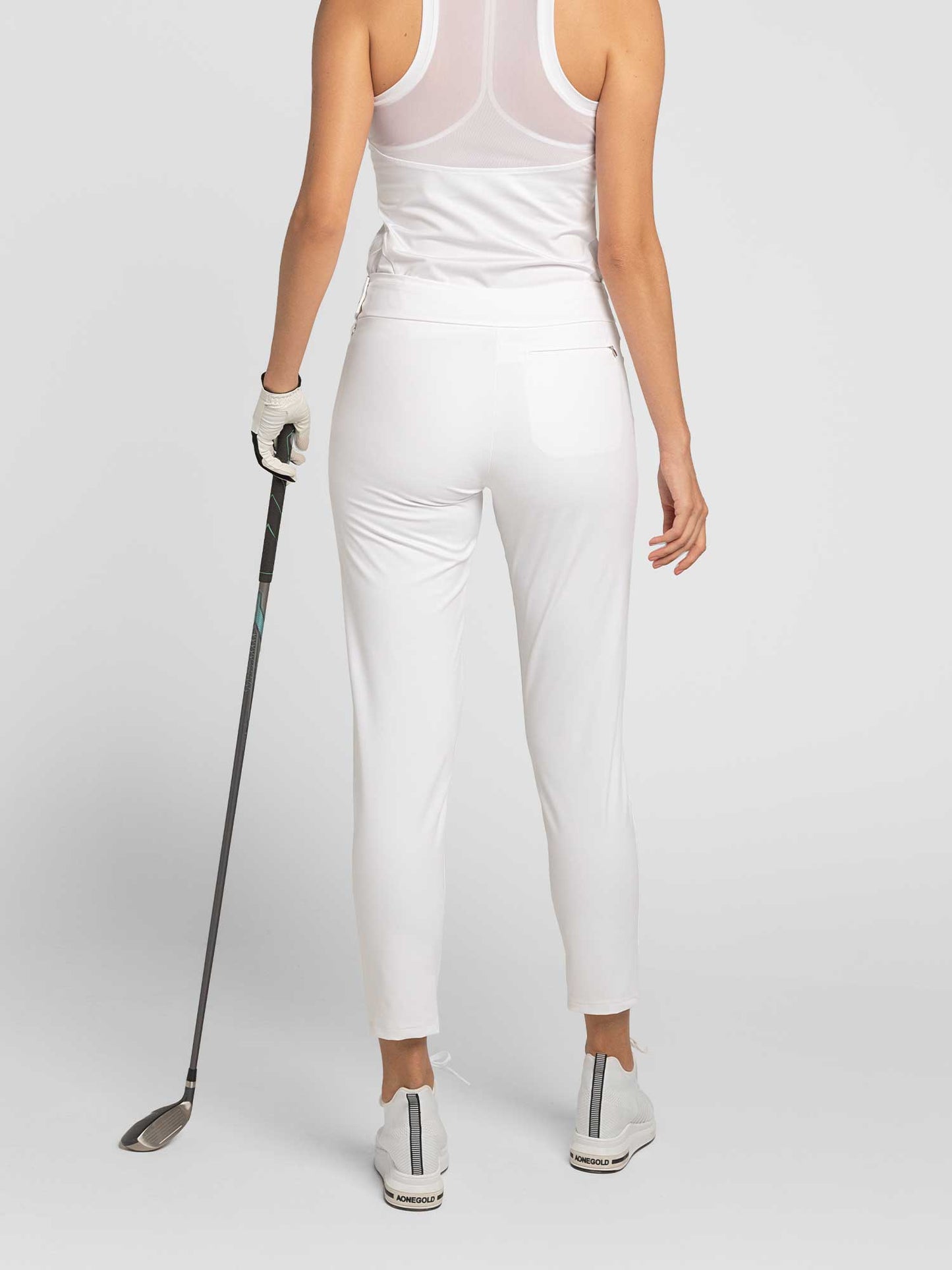 A woman faces away, holding a golf club in her left hand. She wears a white sleeveless top, inPhorm NYC Stylish Women's Golf Pants with moisture-wicking, white golf shoes, and a white glove against a plain light background.