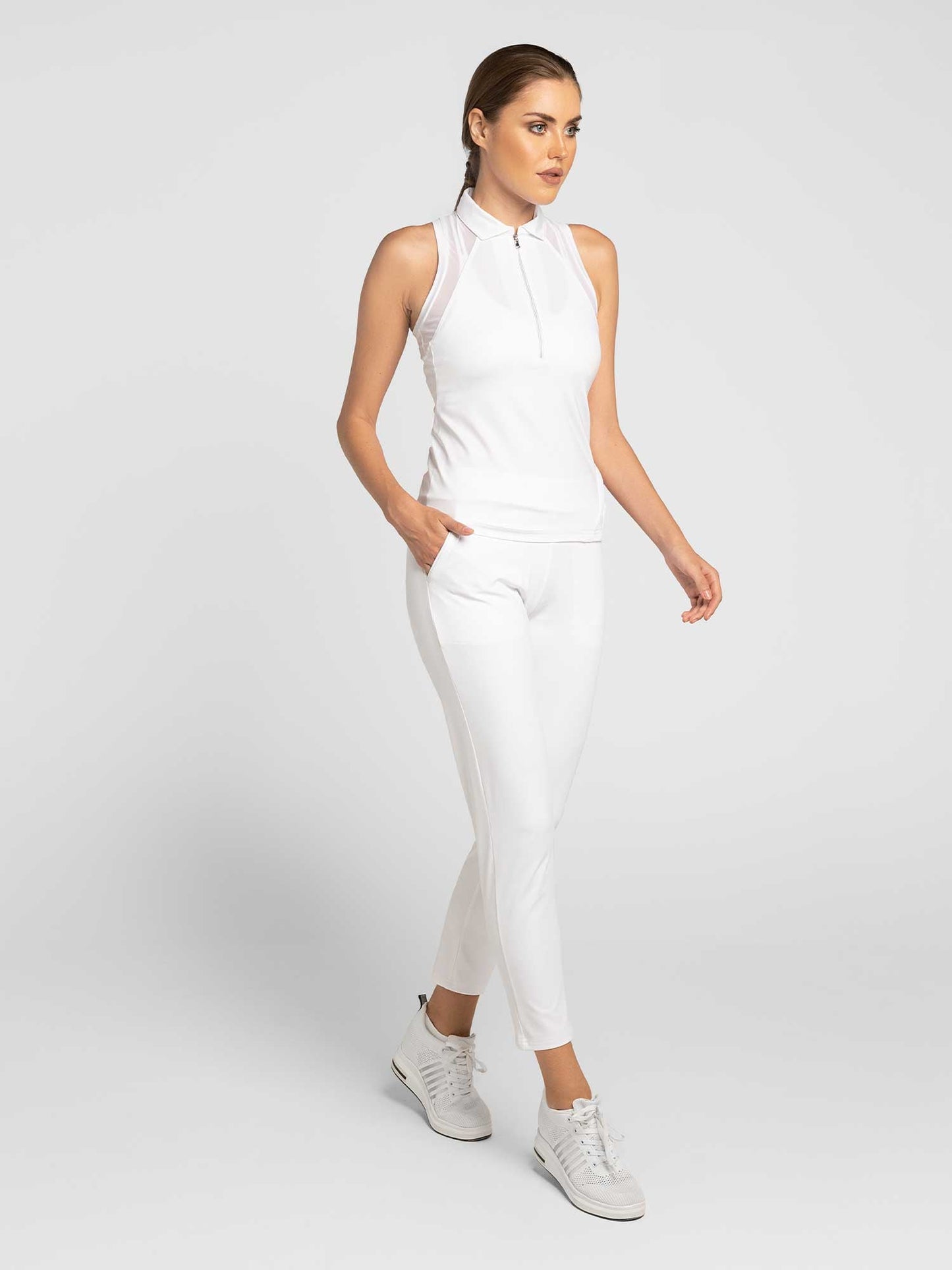 A woman with light skin and brown hair tied back models the inPhorm NYC Stylish Women's Golf Pants—white, fitted, and moisture-wicking—paired with a sleeveless zip-neck top and white sneakers against a plain light-gray background.