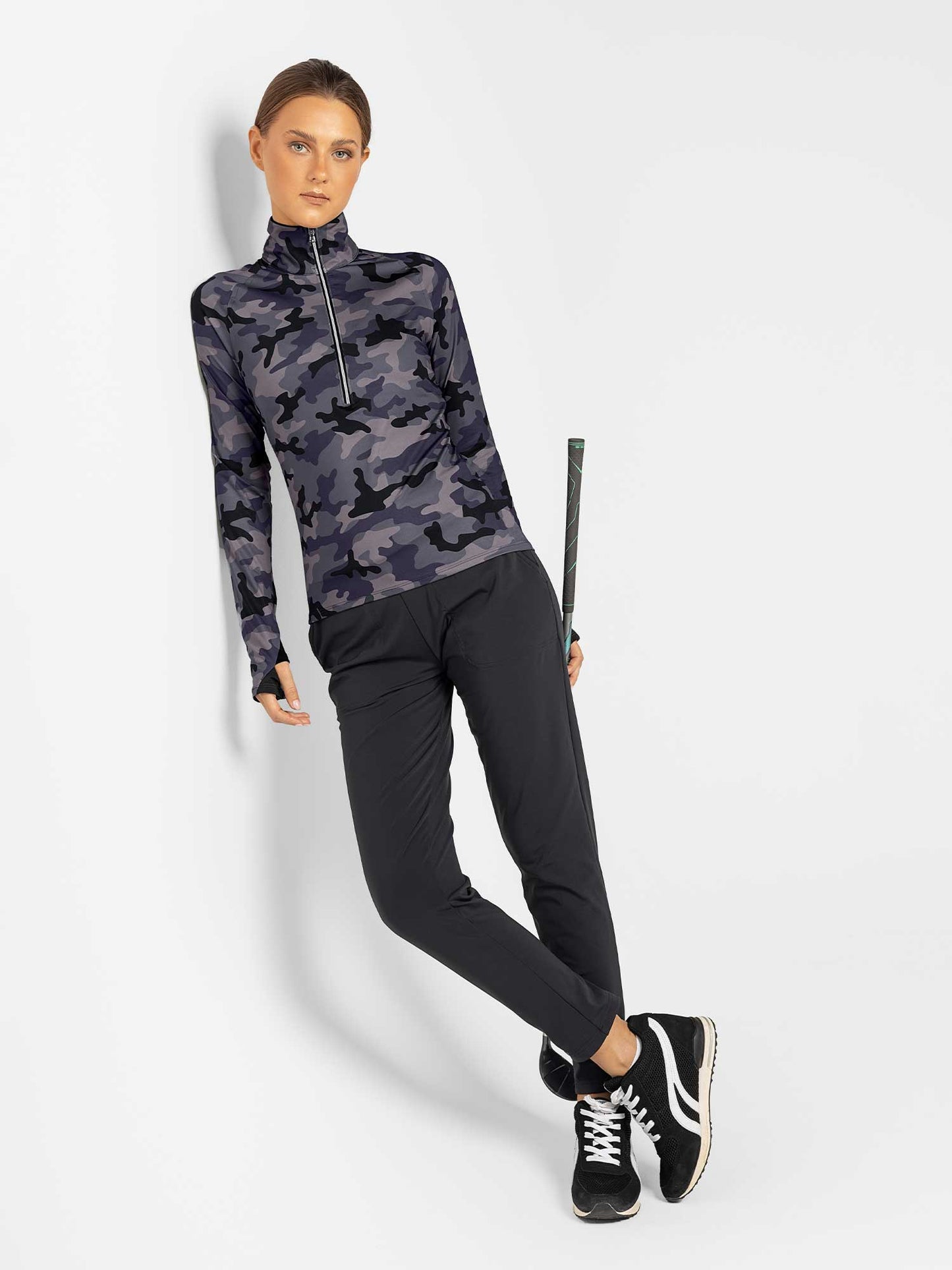 A woman with brown hair pulled back stands against a white wall, holding a tennis racket. She wears inPhorm NYC Stylish Women's Golf Pants—fitted, moisture-wicking black pants—paired with a blue and black camo long-sleeve top and sneakers.
