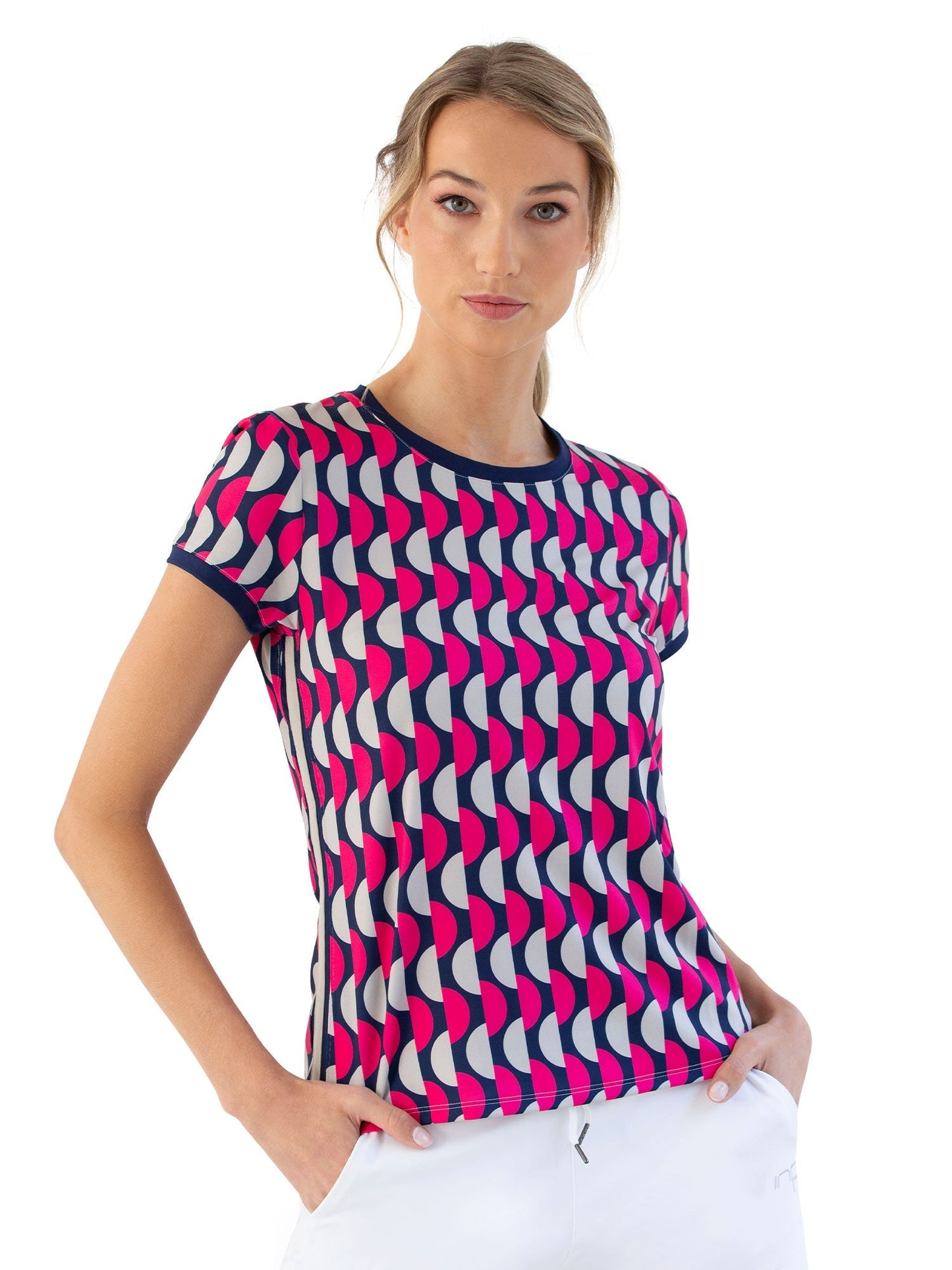 A woman with blonde hair in a loose ponytail wears the inPhorm NYC Sustainble Harper Short Sleeve Crew Neck - Geo Print, featuring bold navy, pink, and white geometric patterns. She stands confidently with her hands in her white pants pockets.