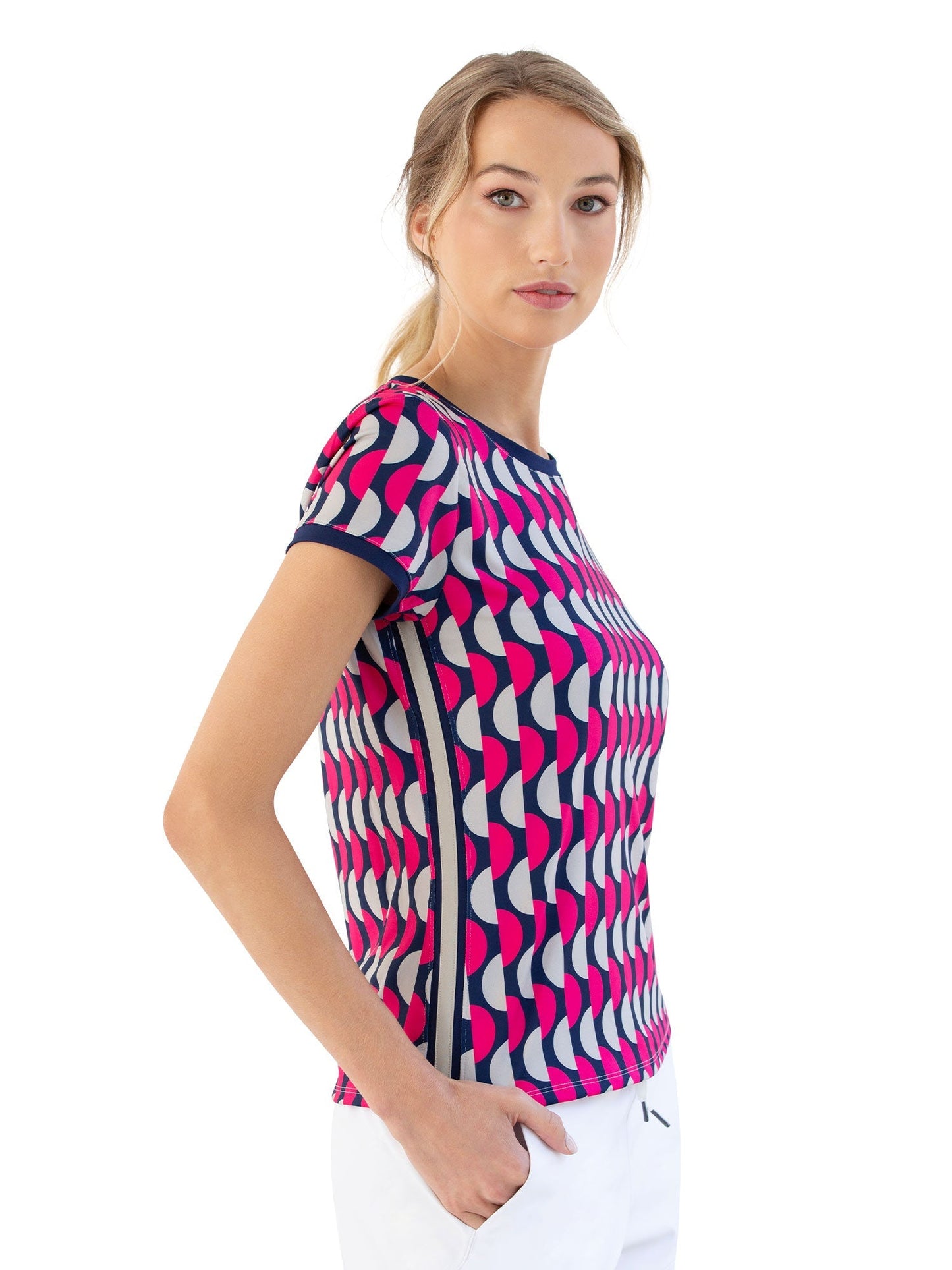 A woman with blonde hair in a low ponytail wears the inPhorm NYC Sustainable Harper Short Sleeve Crew Neck - Geo Print and white pants, standing in profile with her hand in her pocket, looking at the camera against a white background.