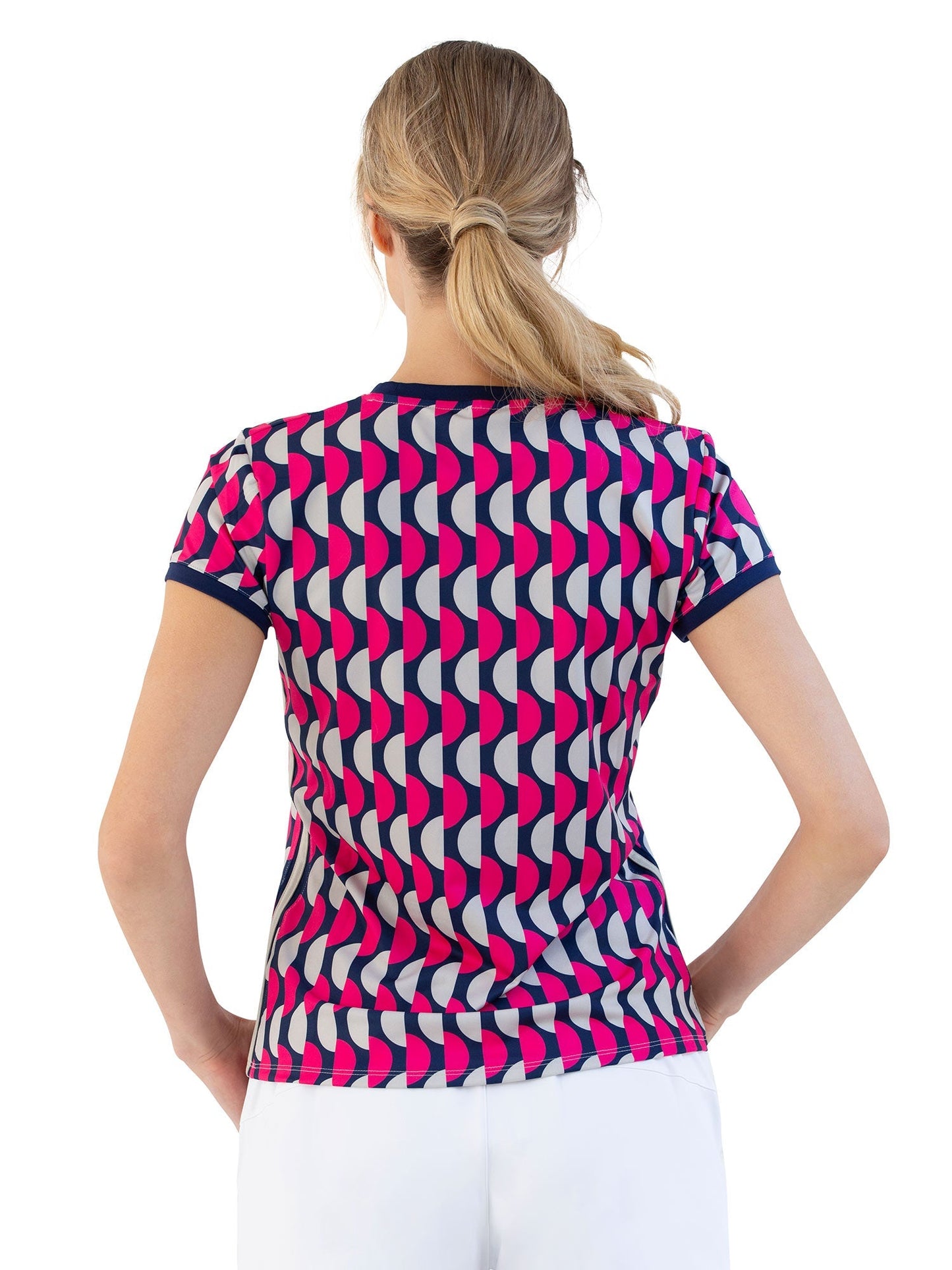A woman with a low ponytail models the inPhorm NYC Sustainable Harper Short Sleeve Crew Neck in Geo Print, featuring a bold pink, navy, and white design. She pairs it with white pants, standing against a plain white background.