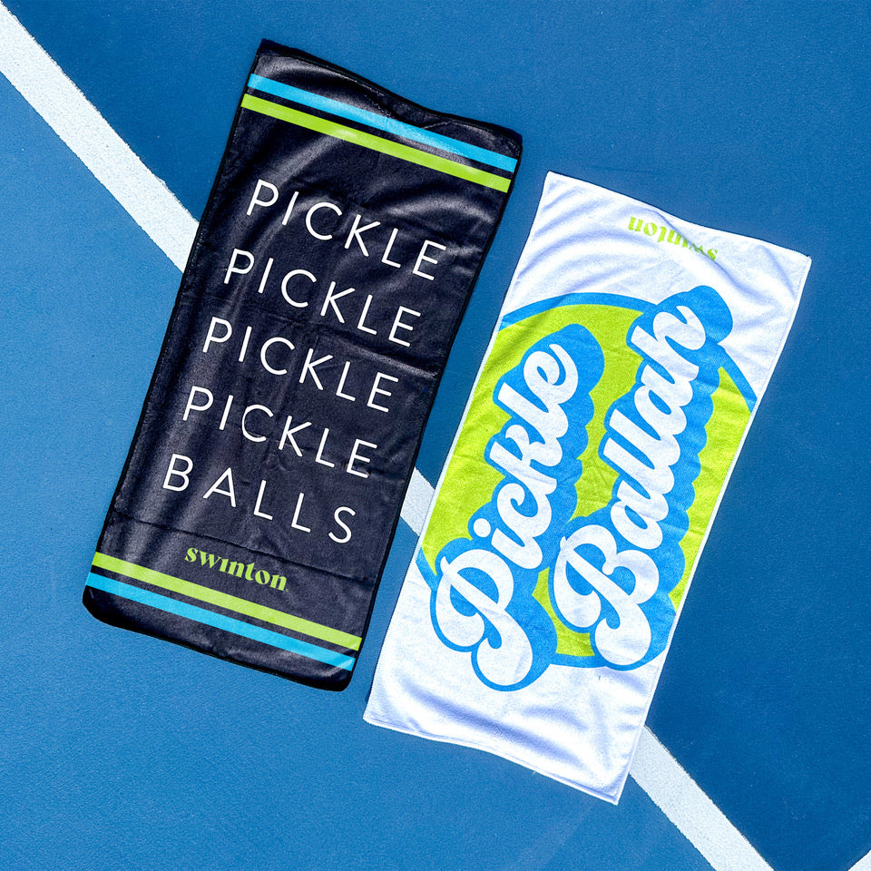 Two rectangular microfiber Swinton Pickleball Sports Towels lie on a blue court. The left is the black Pickleball Sports Towel featuring green and blue stripes and branding; the right is white with "Pickle Ballah" in blue and a green ball.
