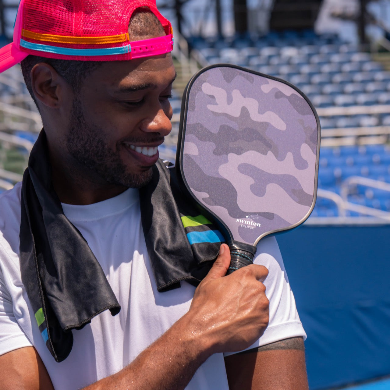 A smiling man in a pink cap and white shirt holds a Swinton Pickleball Eclipse Camo Pickleball Paddle with a textured graphite face. He has a black towel over his shoulders, seated in an empty blue outdoor stadium.