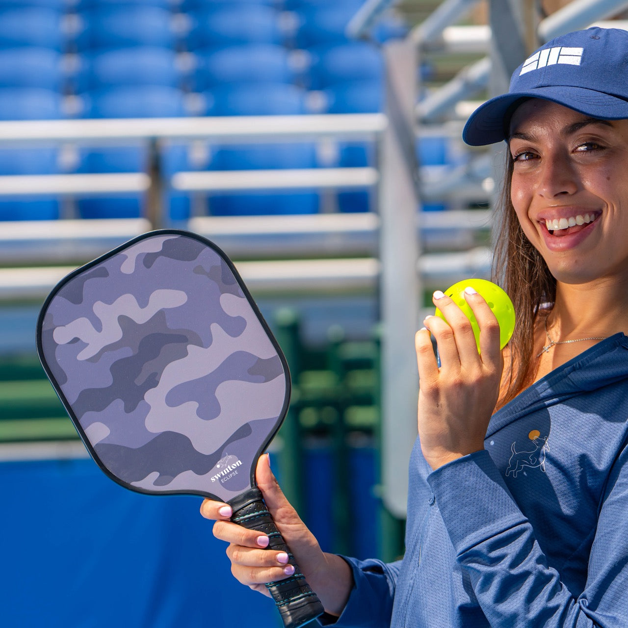 A smiling woman in a navy cap and jacket holds a yellow pickleball and the Swinton Pickleball Eclipse Camo Pickleball Paddle. She stands on an outdoor court with blue stadium seats behind her, sunlight illuminating her face.