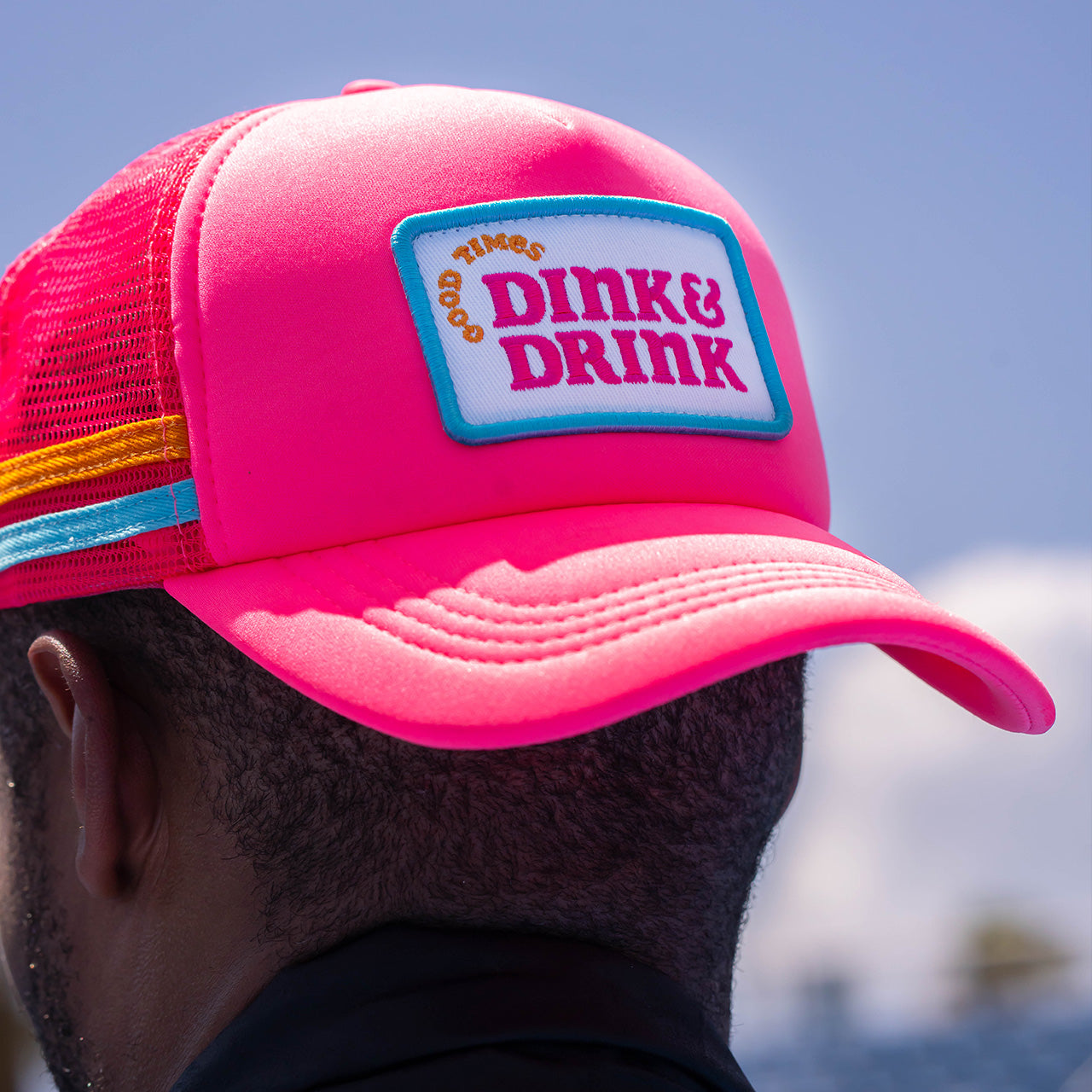 A person wears the Swinton Pickleball Dink & Drink Pink Foam Snapback Hat, featuring colorful stripes and a “GOOD TIMES DINK & DRINK” patch. The adjustable snap closure is visible, with only the side and back of the hat shown against a blue sky.
