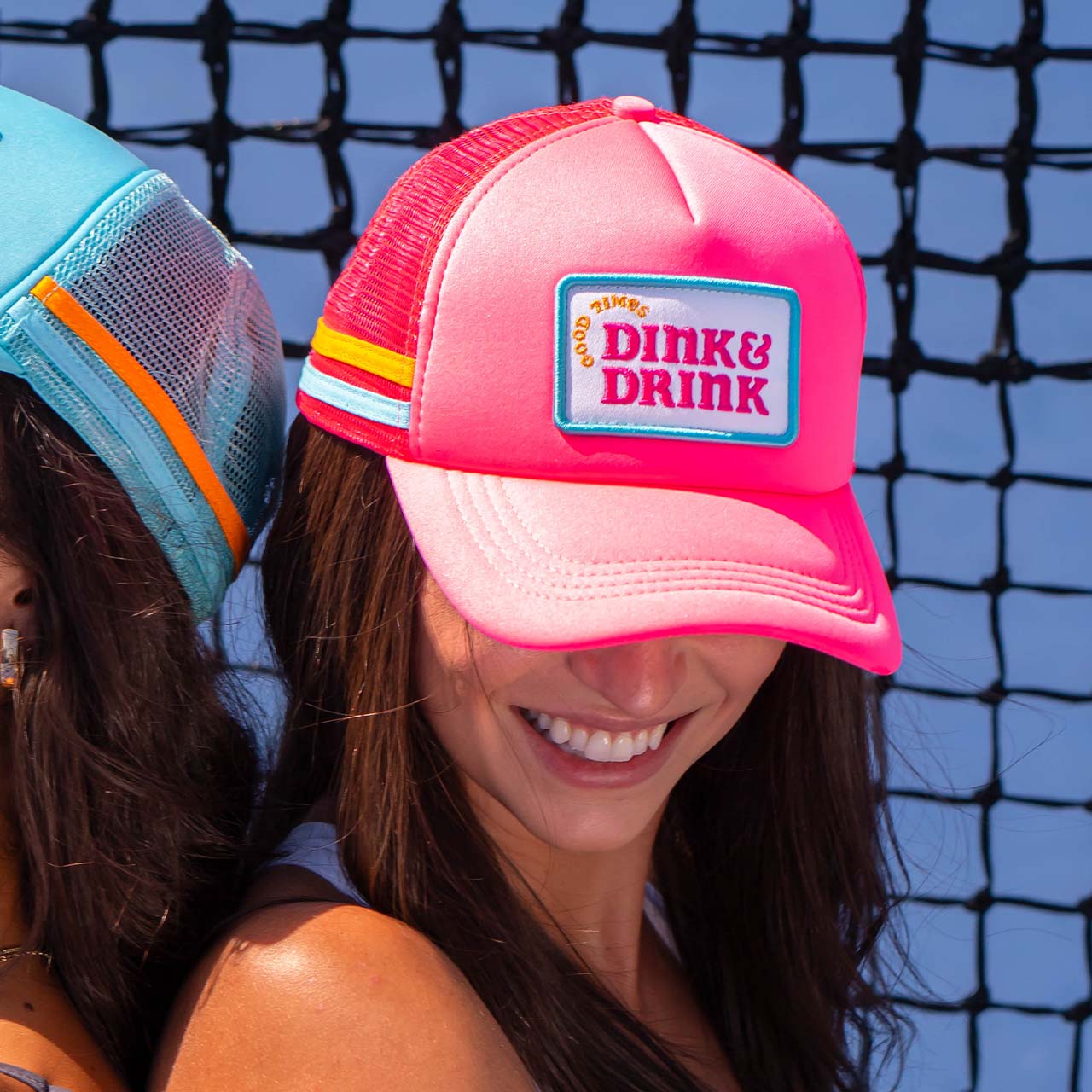A woman smiles in a Swinton Pickleball Dink & Drink Pink Foam Snapback Hat, featuring a rainbow stripe and patch—perfect for pickleball. She stands back-to-back with another woman in a blue cap, with a net and blue sky behind them.