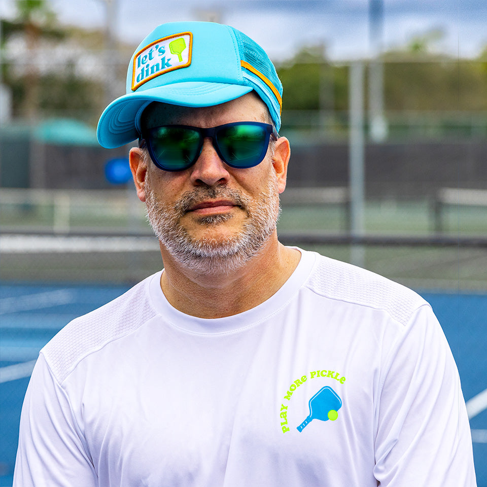 A man with a gray beard wears a turquoise Swinton Pickleball Dinking Cap Foam Snapback Hat, dark sunglasses, and a PLAY MORE PICKLE shirt. He stands on a blue outdoor tennis court with a chain-link fence behind him.