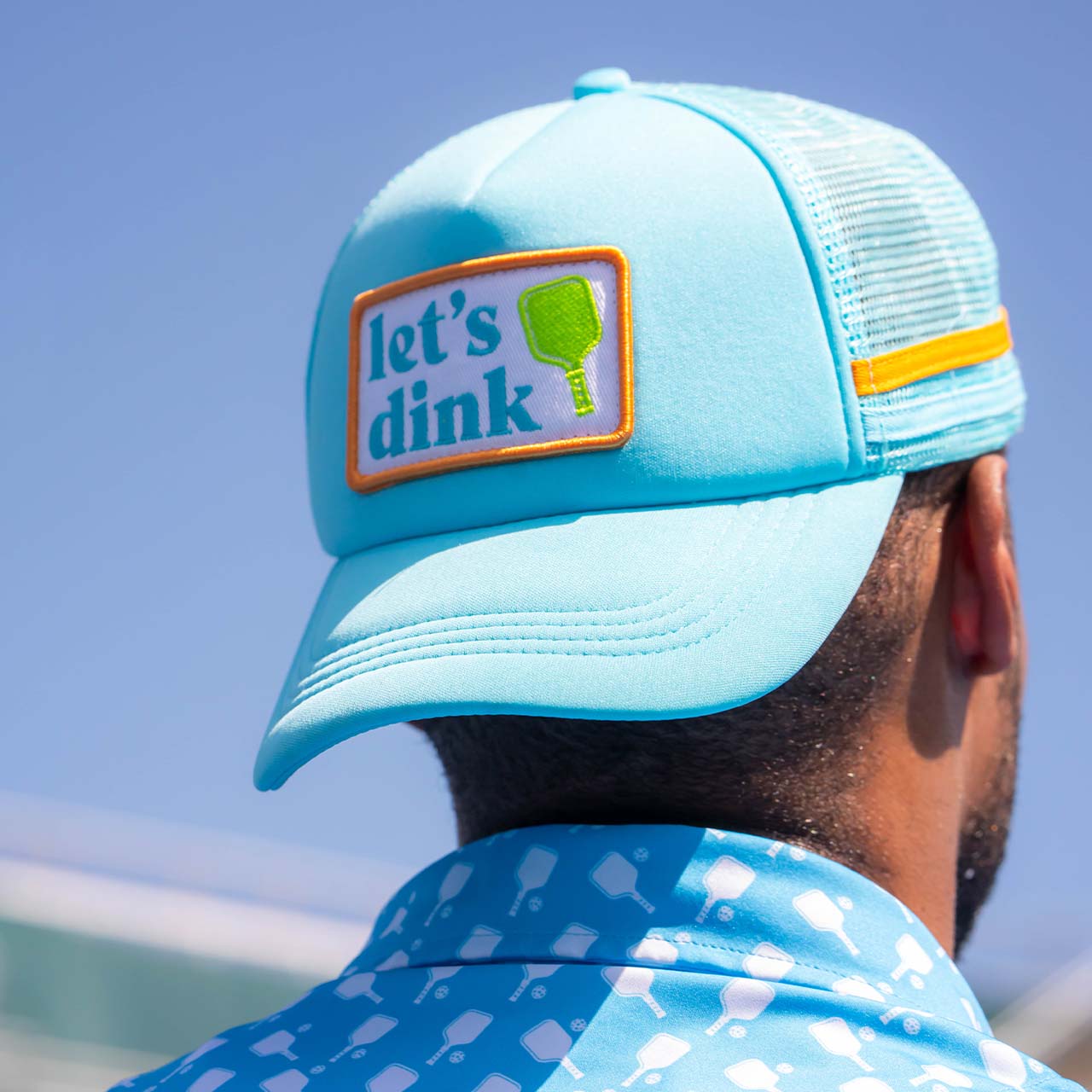 A person wears the Swinton Pickleball Dinking Cap Foam Snapback Hat in light blue mesh, featuring a “let’s dink” patch, and pairs it with a blue geometric shirt against a clear blue sky.
