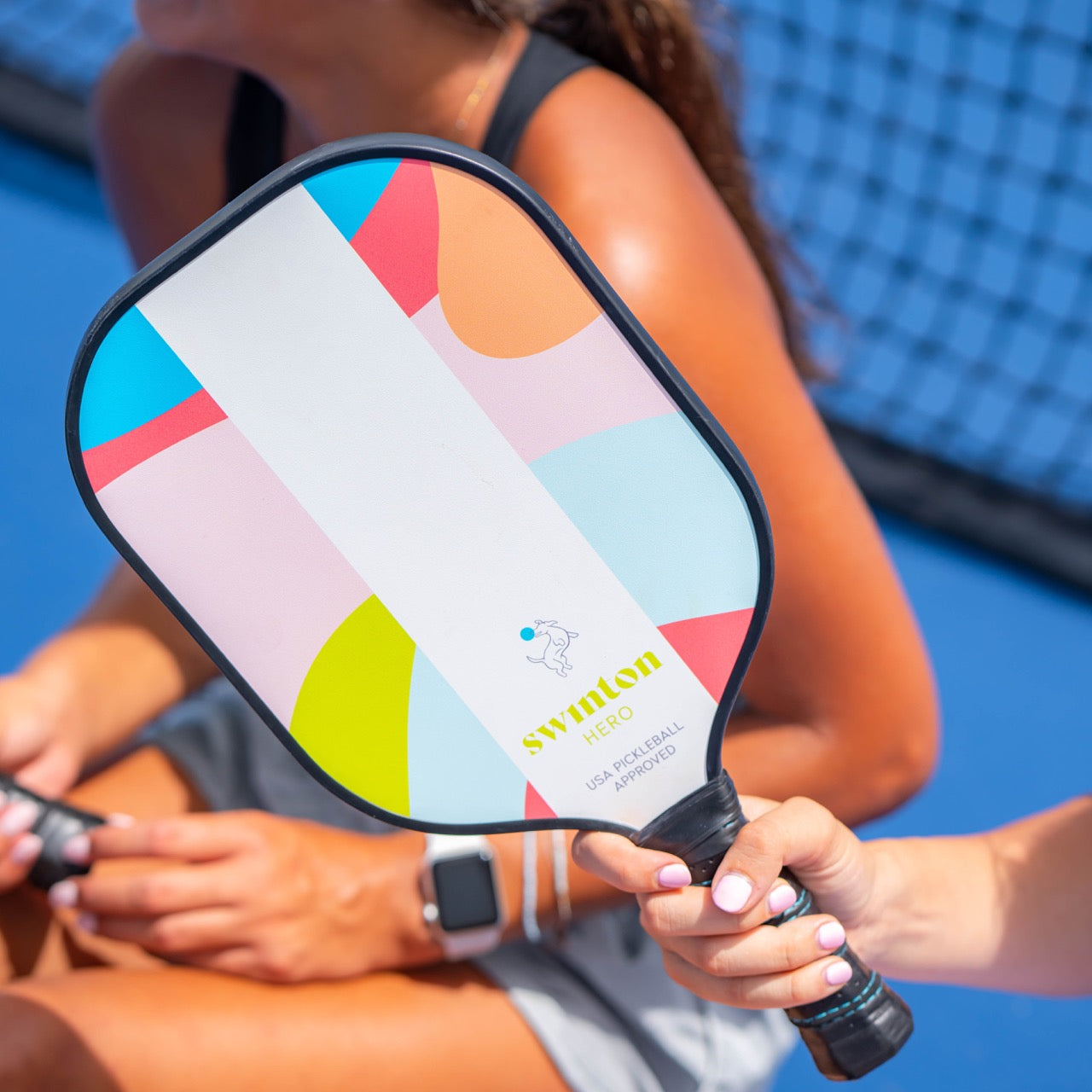 A close-up of a person holding the Hero Breeze Pickleball Paddle by Swinton Pickleball, featuring geometric designs and SWINTON branding. Two women in athletic wear sit on a blue court; one wears a smartwatch. USA Pickleball Approved.