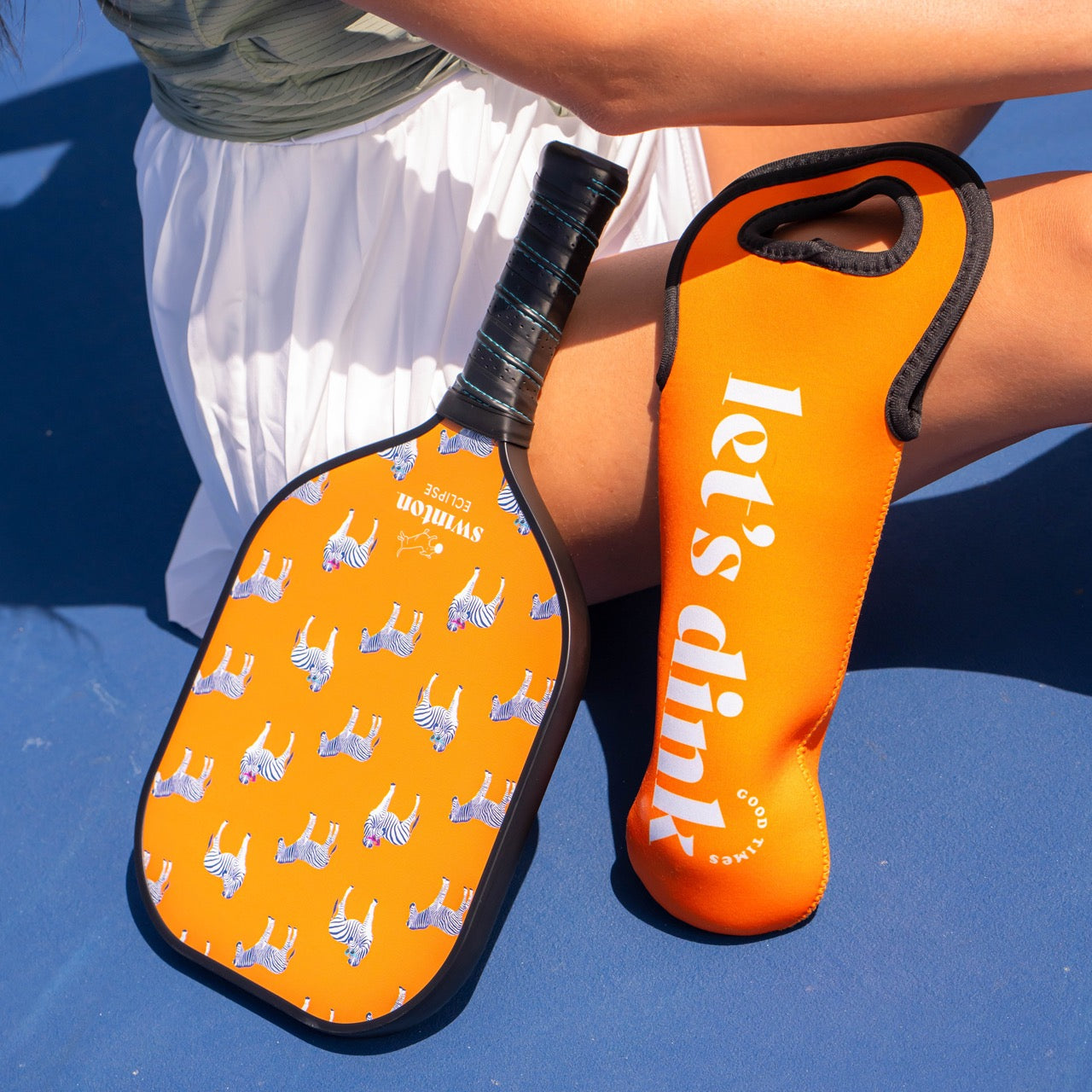 A person in a white skirt sits on a blue court, holding the Swinton Pickleball Eclipse Party Animal Paddle with small blue and white tigers. Beside them is an orange "let’s dink" paddle cover.