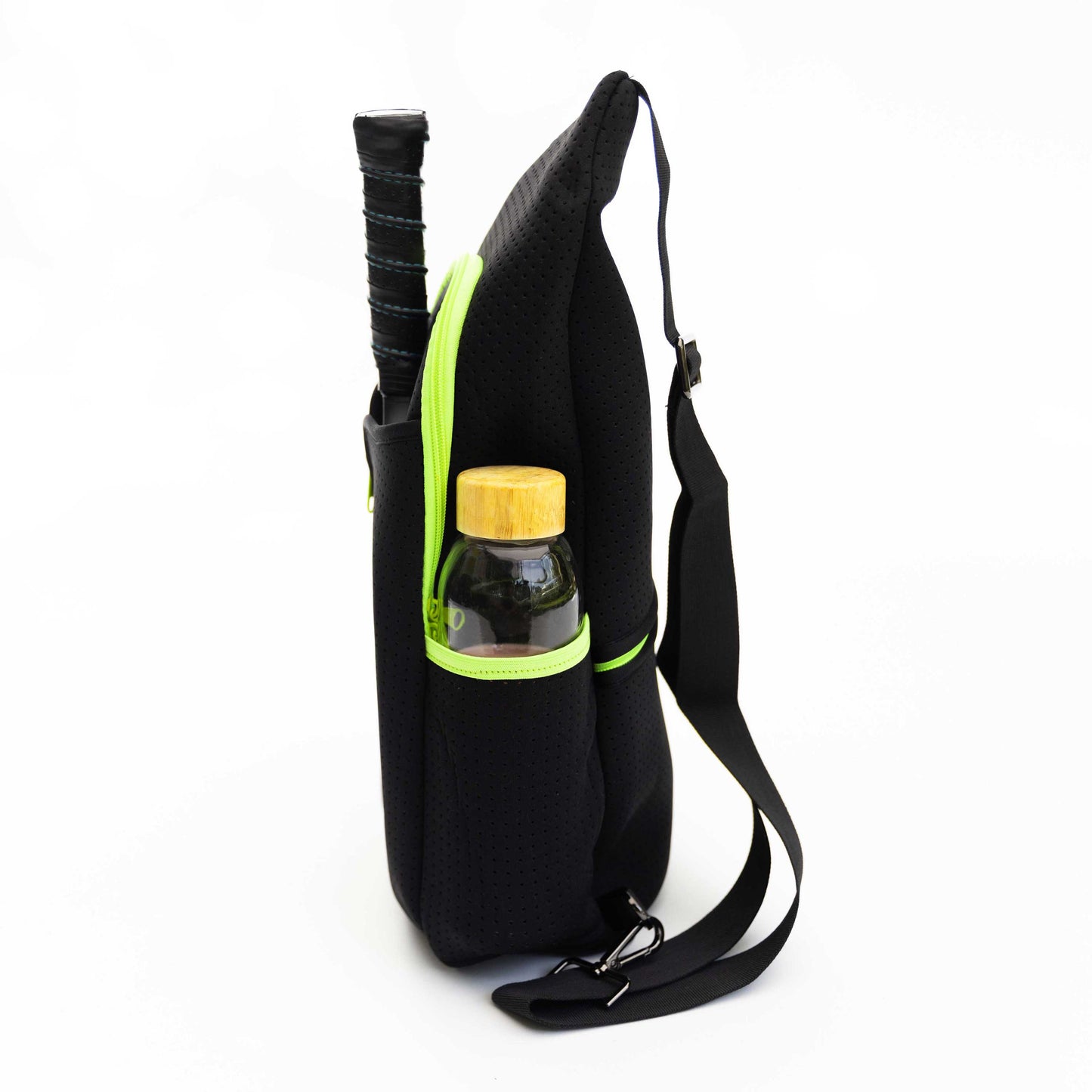 A black Swinton Pickleball Pickleball Sling Bag (personalization available) with a neon green zipper stands upright. A racket handle sticks out of one side pocket; a glass water bottle with wooden lid is in the other. The adjustable strap lies across a white background.