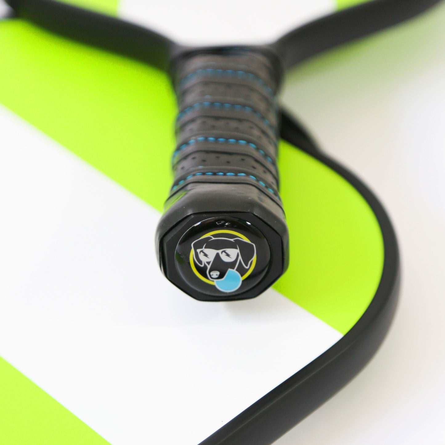 Close-up of the Swinton Pickleball Hero Green Stripe Paddle's black handle with blue stitching and premium grip, dog-in-sunglasses logo with blue droplet on the end cap, and a lightweight face with bright green and white diagonal stripes.