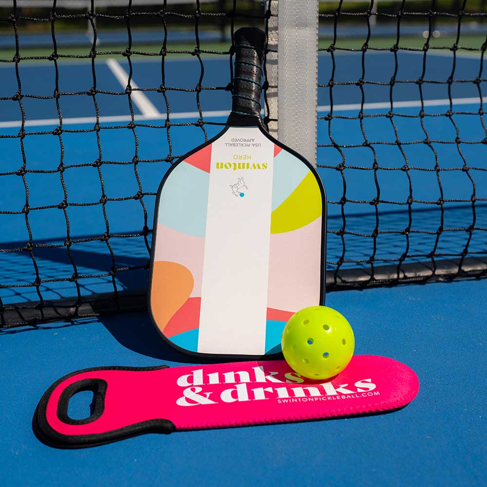 A Swinton Pickleball Hero Breeze Paddle rests by a black net on a blue court, with a yellow pickleball and pink “dinks & drinks” paddle cover in front. Sunlight casts shadows on this USA Pickleball Approved setup.