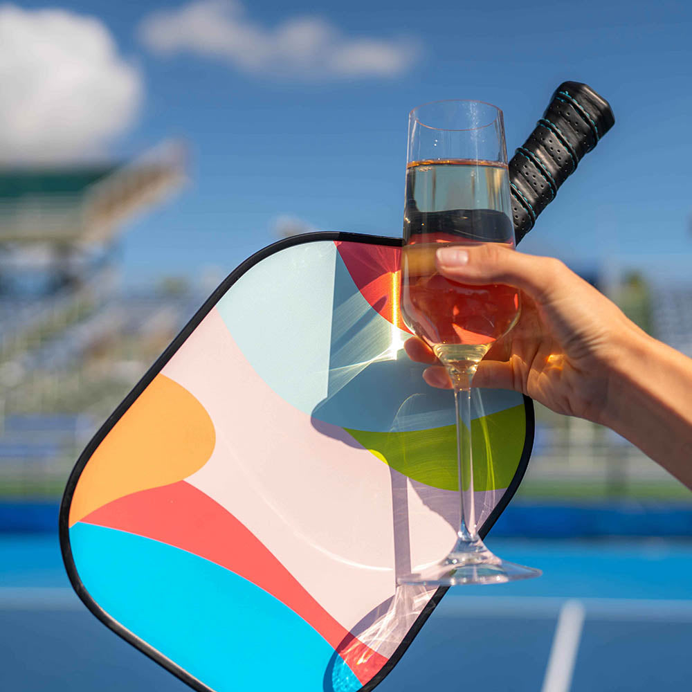 A hand holds a glass of rosé wine in front of the Swinton Pickleball Hero Breeze Paddle, with the graphite paddle resting on a blue outdoor court. Stadium seating and a partly cloudy sky appear blurred in the background.