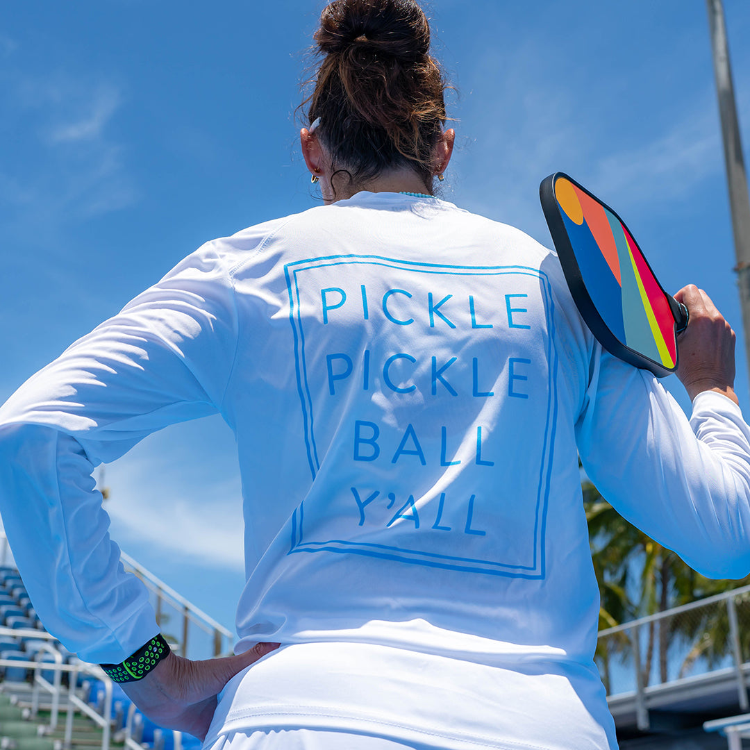 A person stands on a pickleball court holding a colorful paddle, wearing Swinton Pickleball’s white “Pickle Pickle Ball Y'all” UPF 50+ long sleeve water shirt. The clear sky and palm trees form the backdrop.