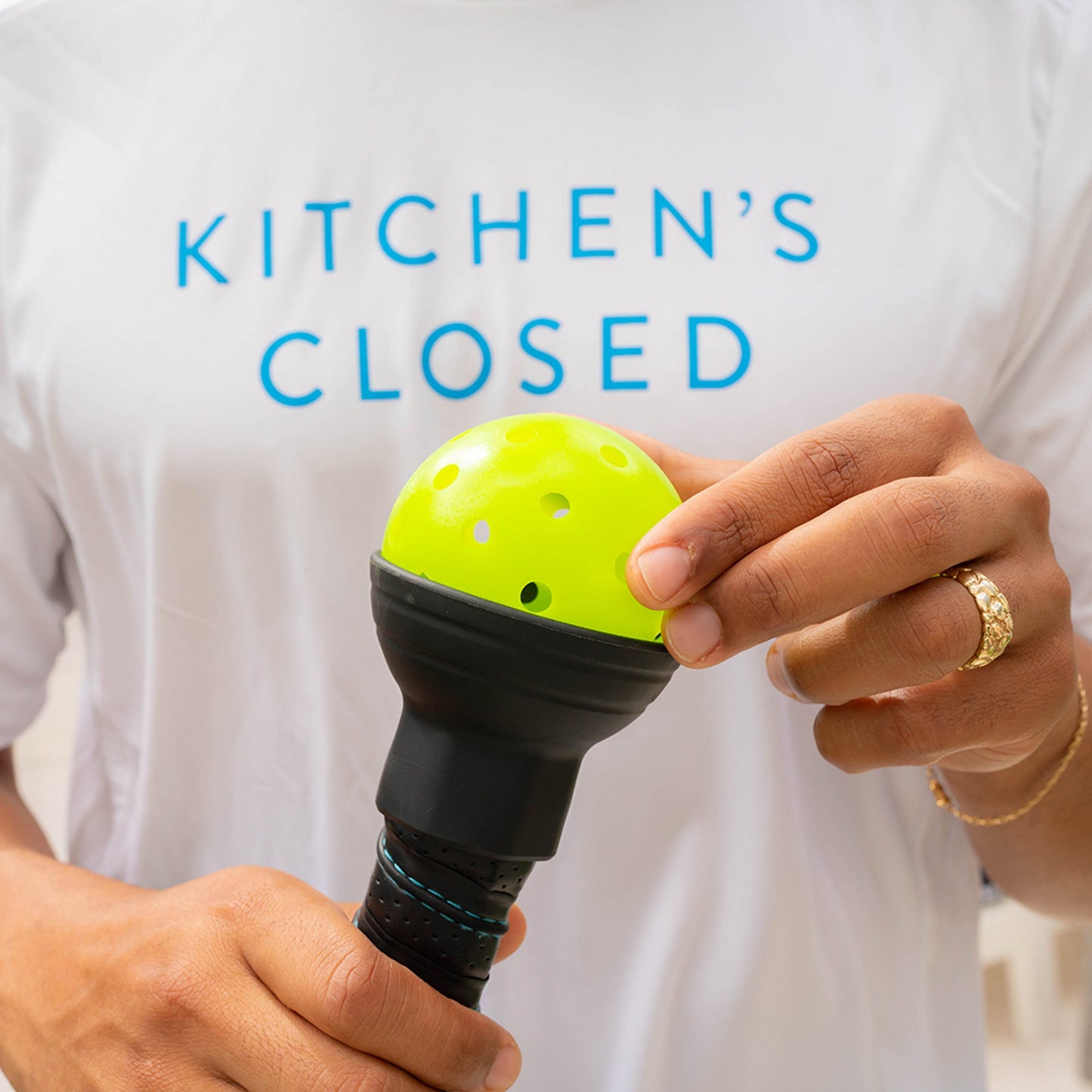 A person wearing a white shirt with blue text reading KITCHENS CLOSED holds the Swinton Pickleball Suck Up, a black paddle with a bright yellow, perforated picker; only their hands and torso are visible, and they wear a gold ring and bracelet.