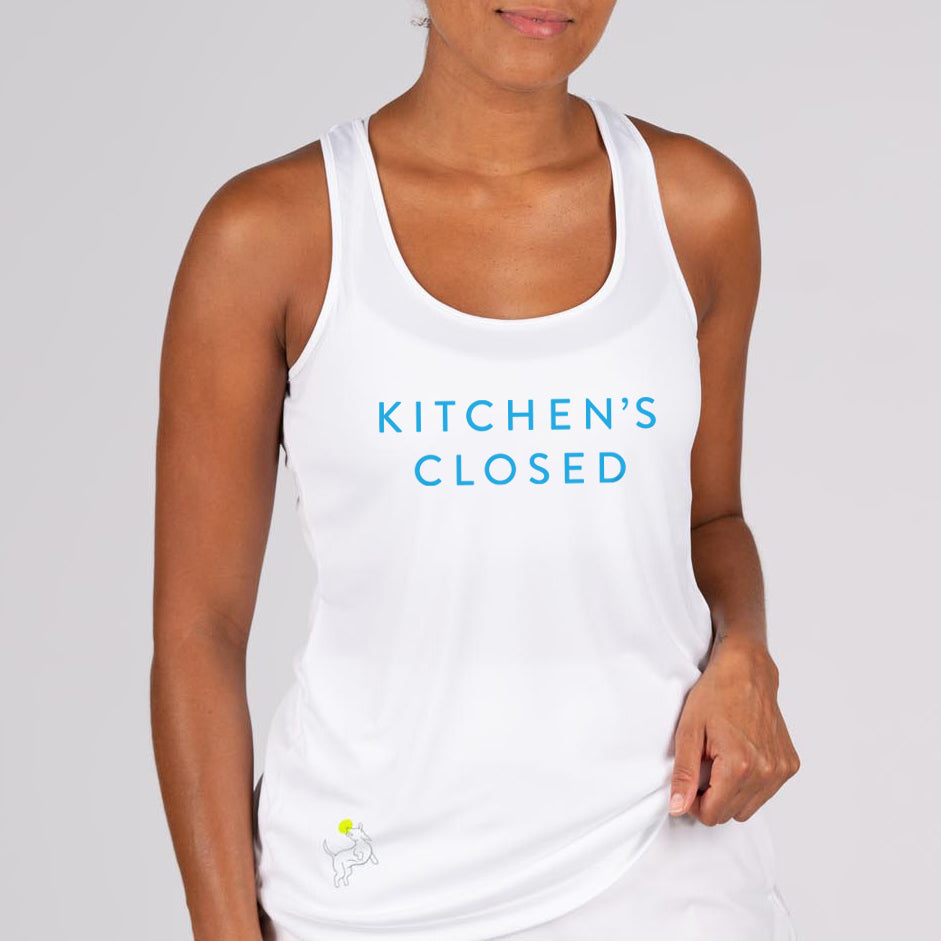 A woman models the Swinton Pickleball Performance Tank White, featuring “KITCHEN’S CLOSED” in bold blue and a yellow tennis ball graphic at the bottom left. She holds the hem against a plain light gray background.