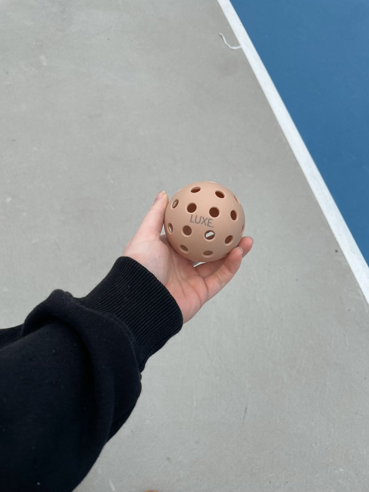 A person in a black sleeve holds a LUXE Pickleball Tan Pickleball (from the 3-pack) with 40 holes above the blue outdoor court and white boundary line.