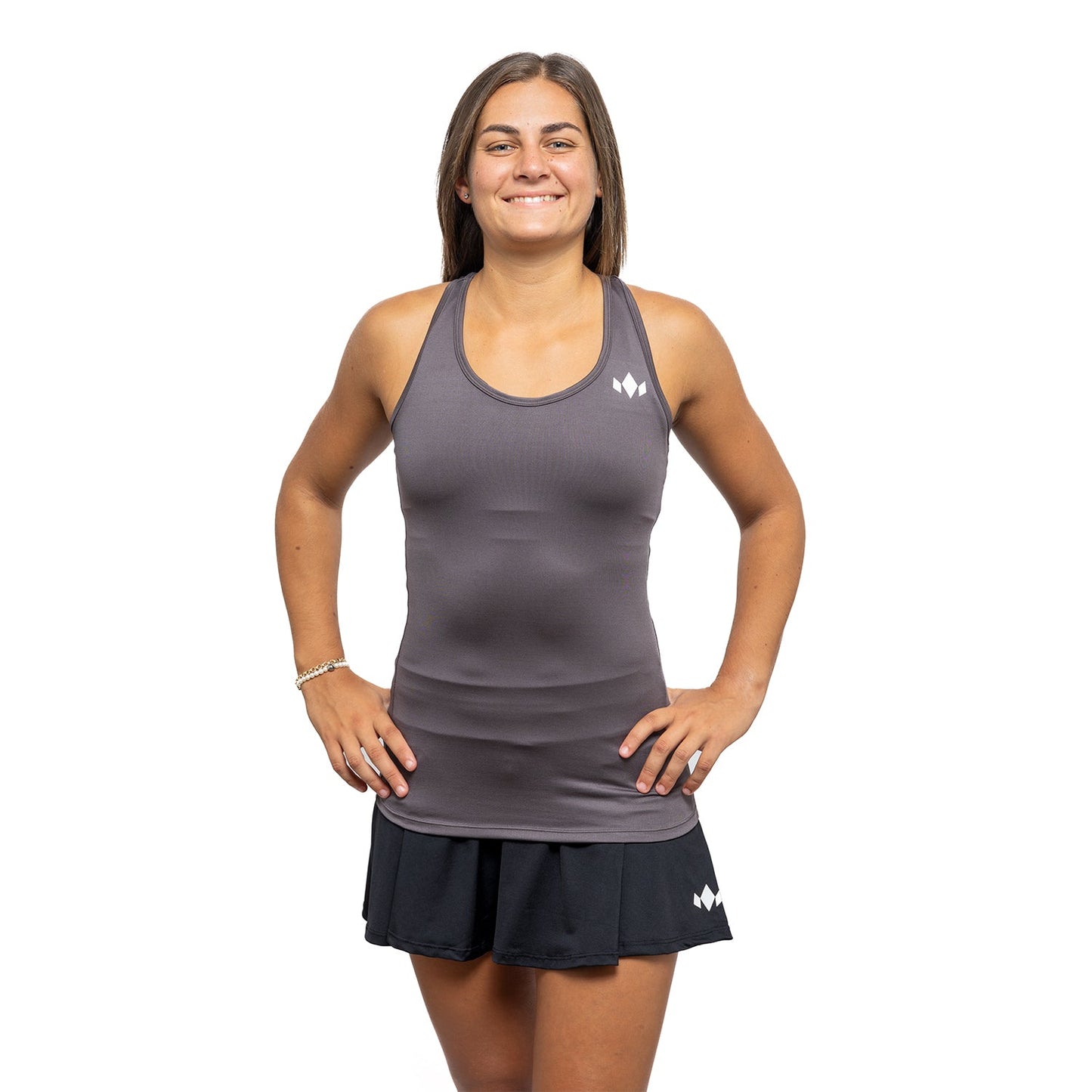 A young woman with long brown hair smiles, standing with hands on hips. She wears the Diadem Sports Team Essential Racerback Tank in gray and a black athletic skirt with white logos, posed against a plain white background.