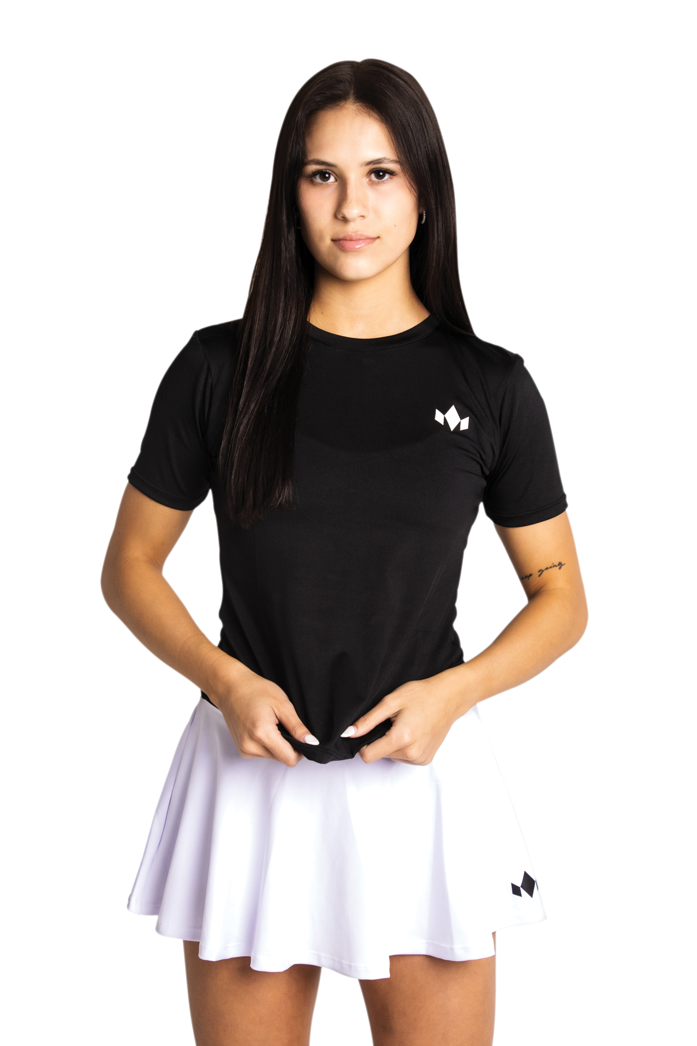 A young woman with straight, dark hair stands facing forward against a plain light background. She wears the Diadem Sports Team Women's Essential Crewneck in black with a white logo, paired with a white tennis skirt and holds her shirt’s hem.