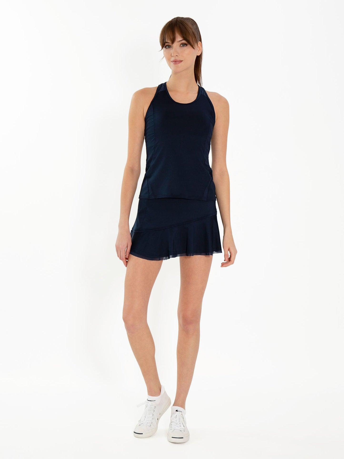 A woman models the inPhorm NYC Tennis Classic 13 1/2" Flounce Skirt in Midnight against a white background, paired with a navy athletic tank, white sneakers, and has straight brown hair with bangs and a neutral expression.