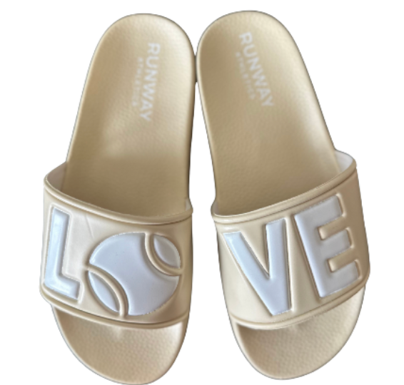 The Tennis LOVE After Play Tennis Slides by Runway Athletics feature beige straps with “LOVE”—the “O” as a tennis ball—and white brand lettering on the insole. A unique tennis gift, these natural and white sandals rest on a clean backdrop.
