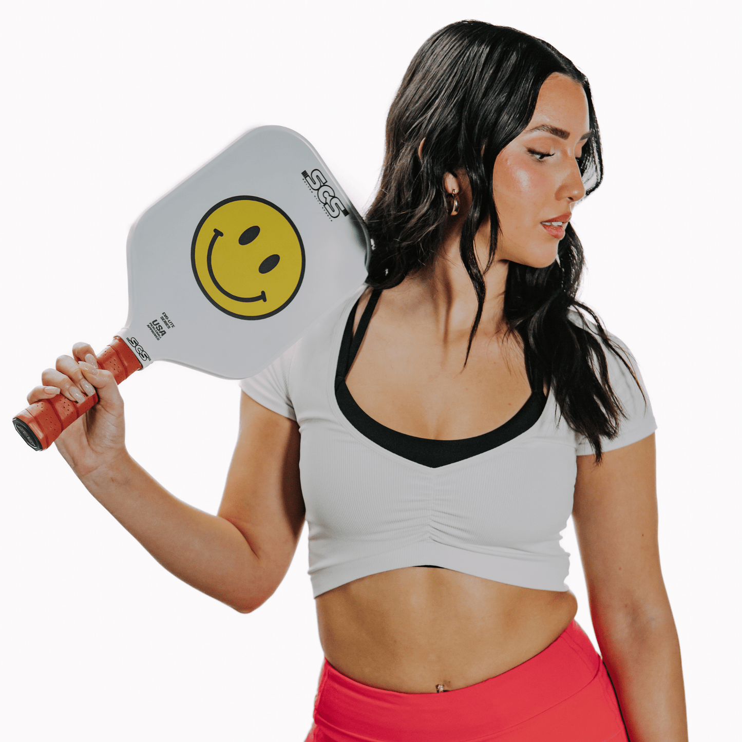 A woman with long dark hair wears a white crop top and pink pants, holding The Happy Paddle | Evo-Lite Series by Salted City Sports, featuring a large yellow smiley face logo, over her shoulder. She stands relaxed against a plain white background.